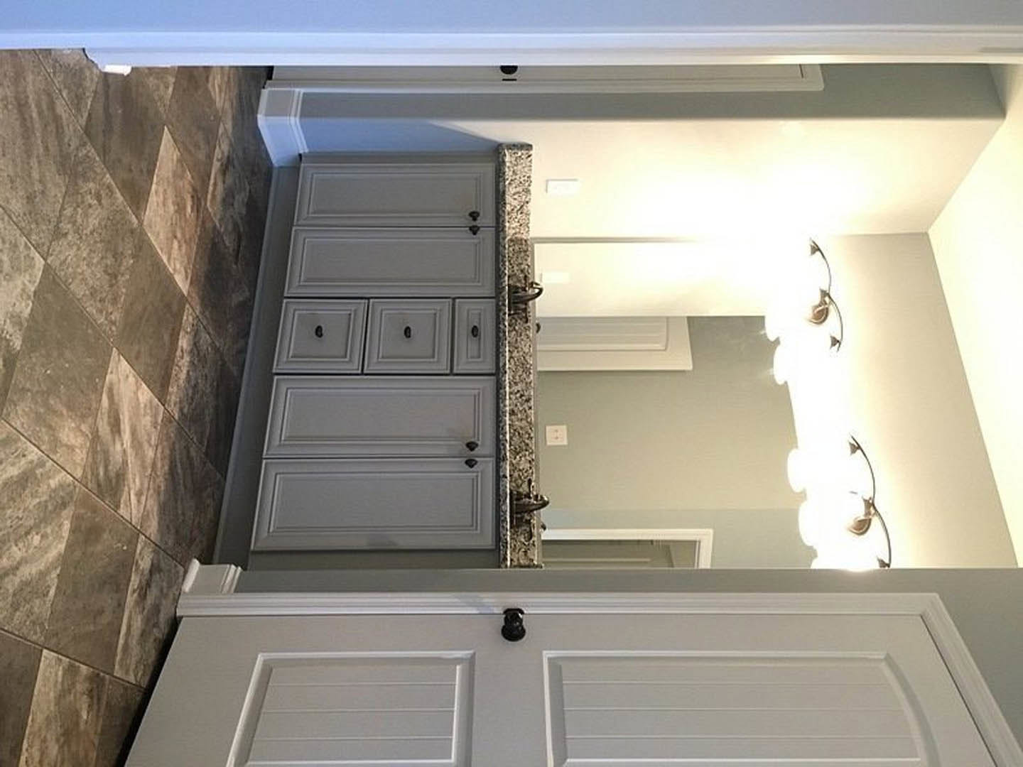Bathroom with white vanity drawers and black handles, rectangular mirror above sink, tile flooring, white walls, and a door-mounted camera visible.