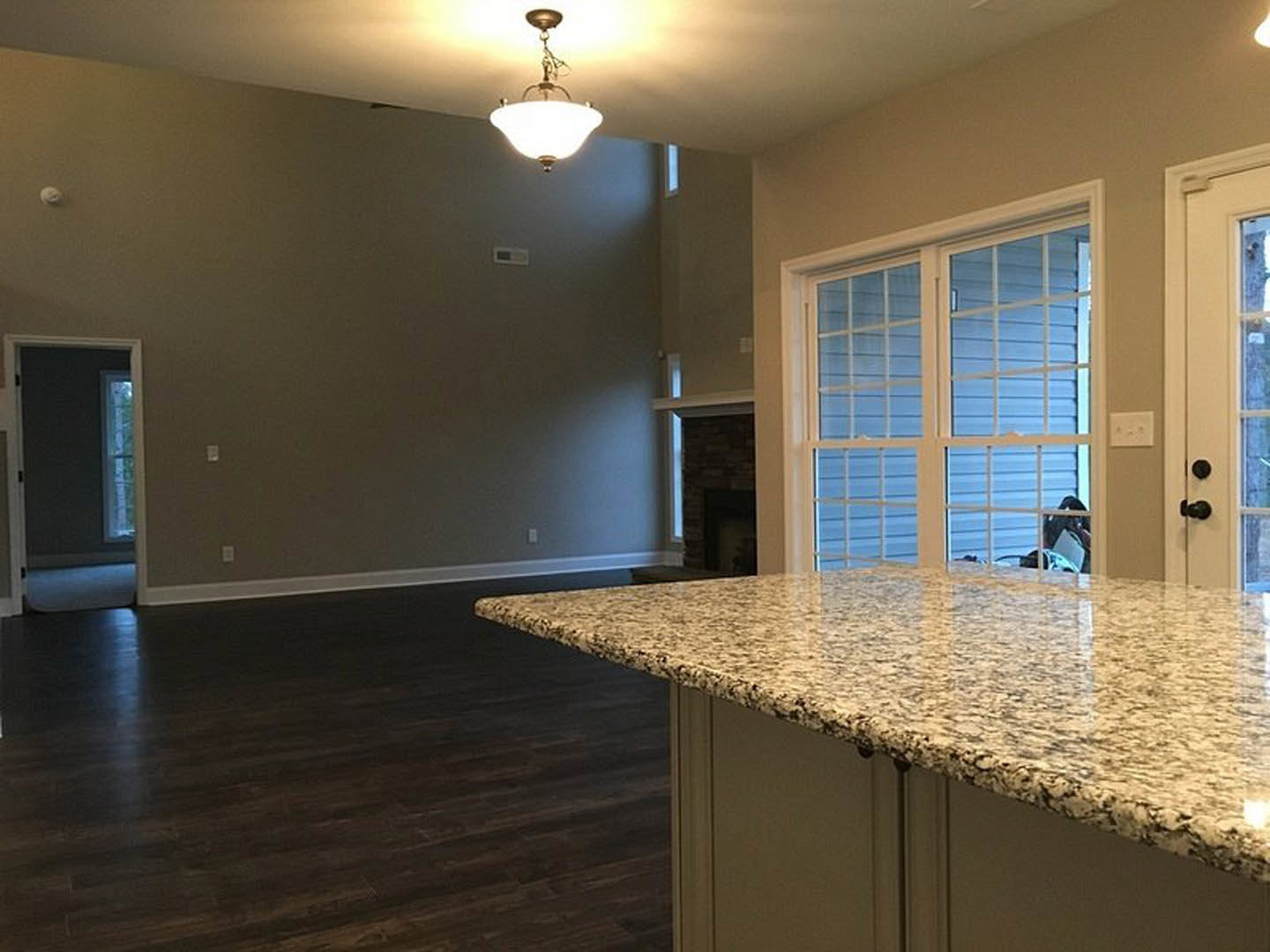 Open-concept kitchen and living room featuring a stone fireplace, granite countertop, white cabinetry, tile flooring, pendant light with white shade, and large windows with glass