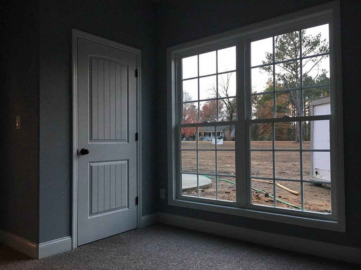 White door with black frame next to a window overlooking a dirt field and trees, beige carpet flooring, metal fence and green-white pole visible outside.