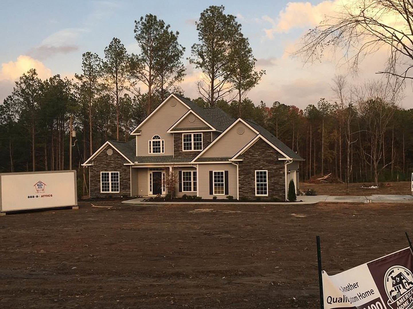 Two-story house with white siding, multiple-paned windows, surrounded by dirt and mature trees under a partly cloudy sky