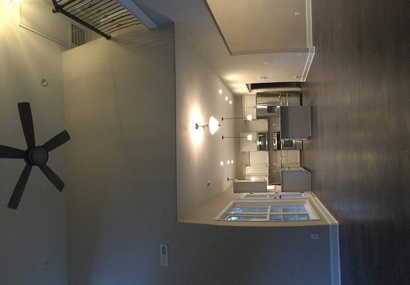 Open-concept kitchen with white cabinetry, stainless steel appliances, wood staircase, ceiling fan with circular blades, wall-mounted light fixture, person standing near a window