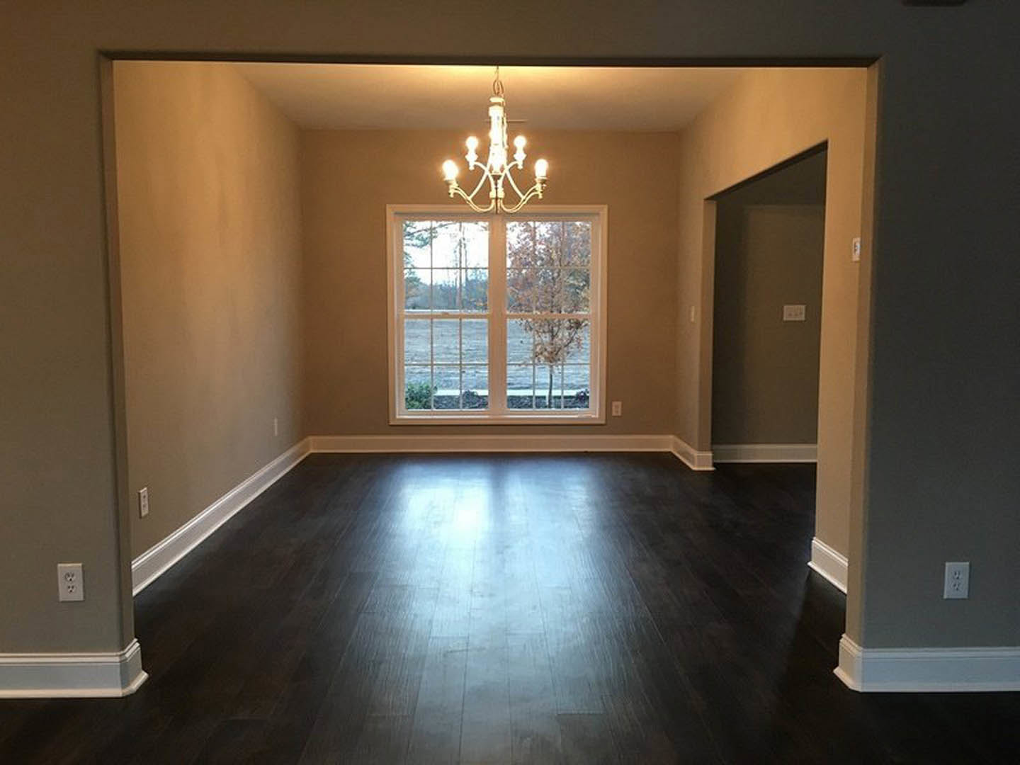 Chandelier with multiple lights hanging from ceiling, large window framed in white molding showing trees outside, wood laminate flooring, light switch on plaster wall