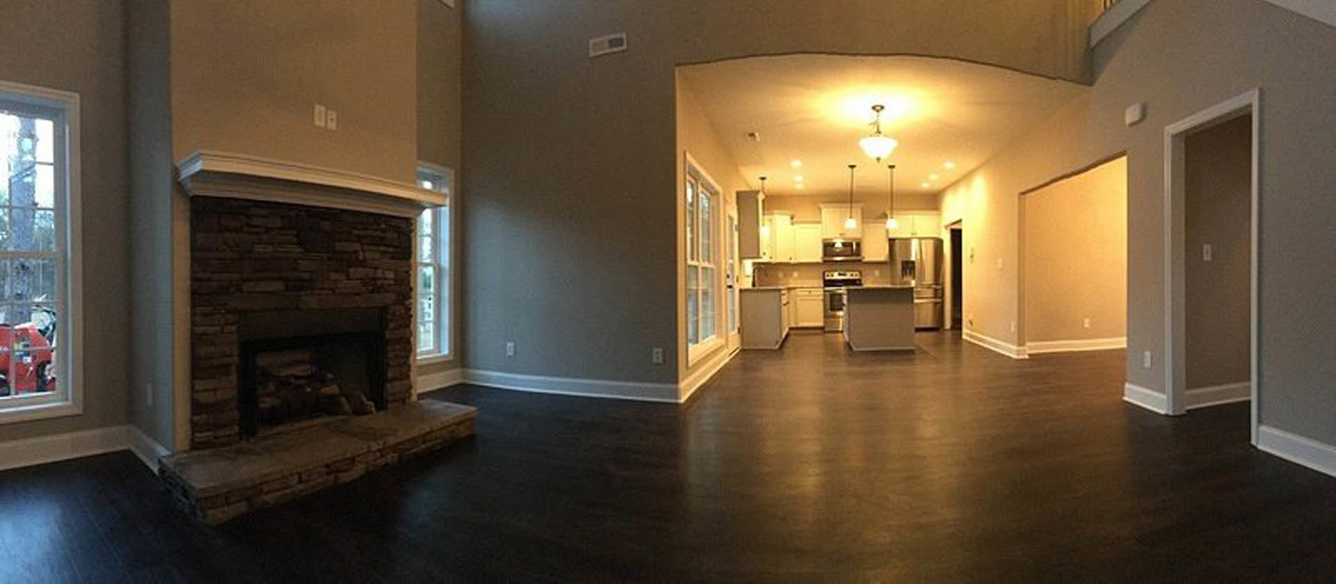 Open concept living room with wood flooring, stone fireplace, and adjacent kitchen featuring white cabinetry and stainless steel appliances