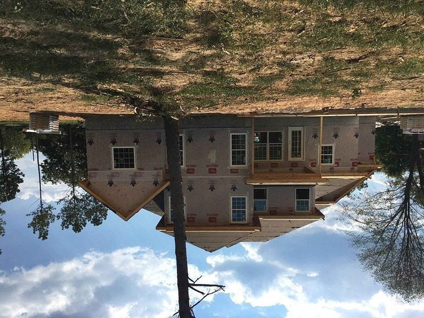 Partially built house with exposed framing, large grid windows, and a mature tree in front, set on a dirt and grass lot under a cloudy sky