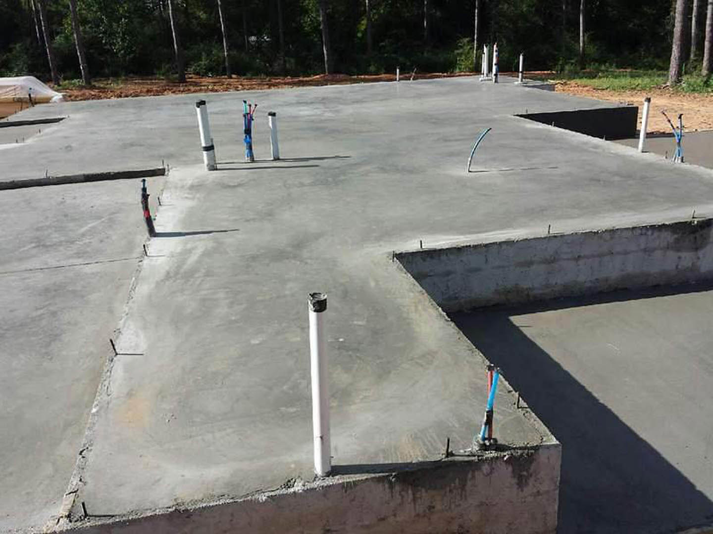 Concrete foundation slab with exposed plumbing pipes and a white pole, surrounded by outdoor construction materials and trees in the background