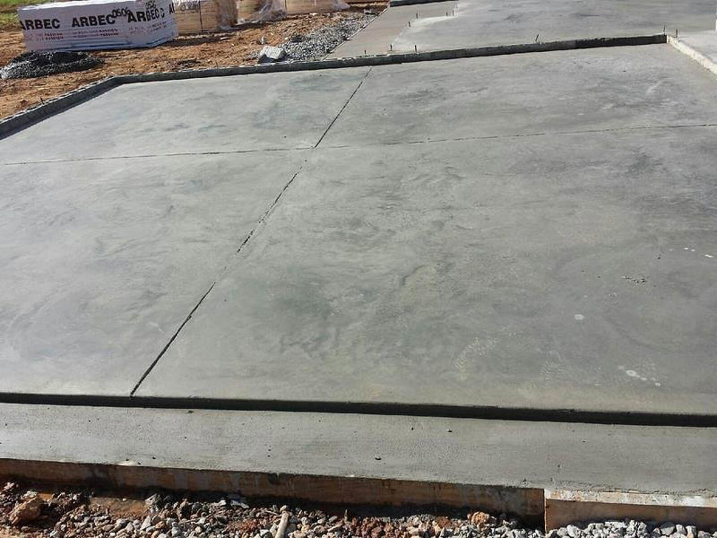 Freshly poured concrete slab with square pattern at residential construction site, bordered by curb and surrounded by gravel and rocks, outdoor signage visible in background