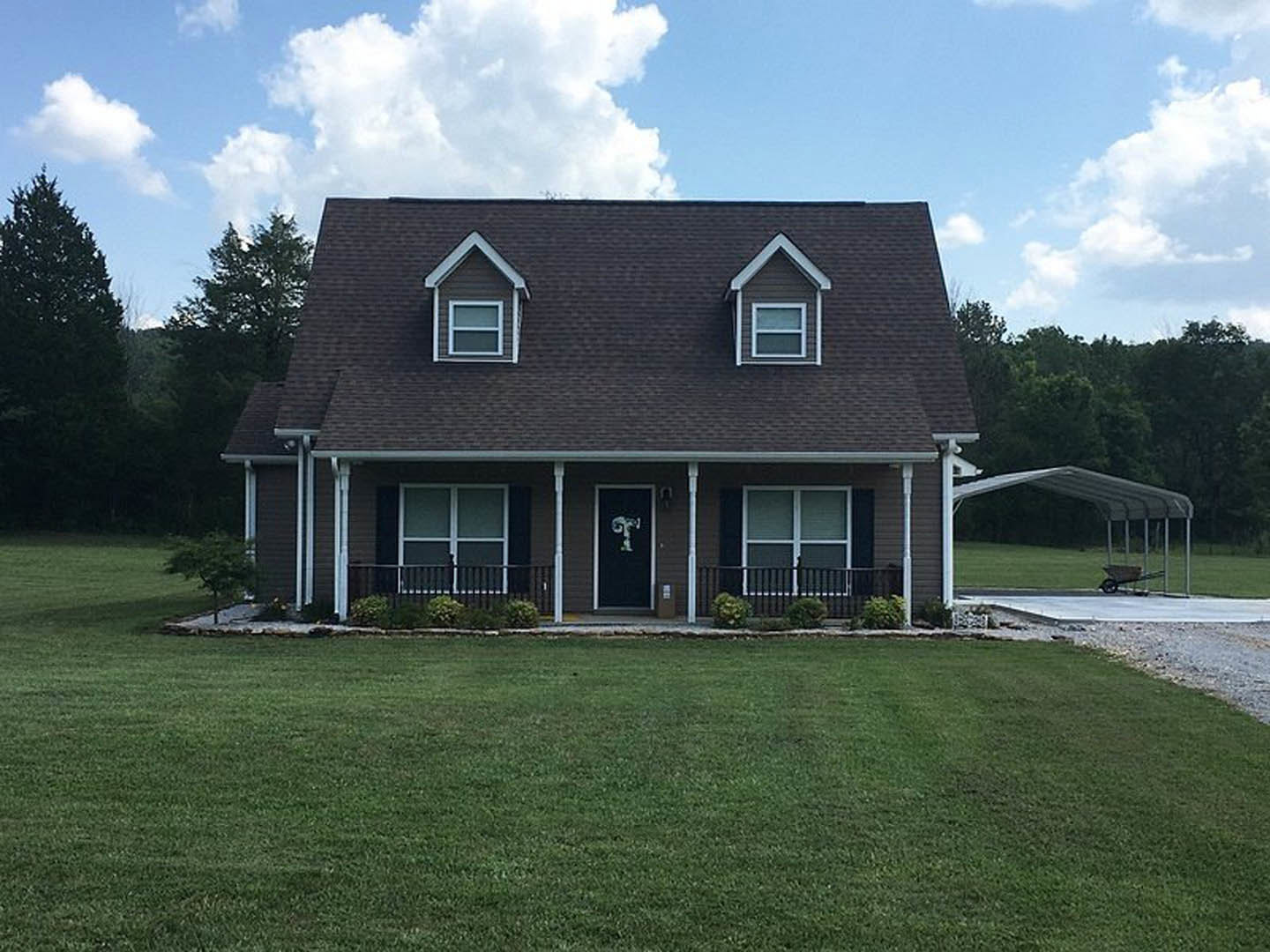 Two-story house with covered porch, black door and railing, white-framed windows, green lawn, wheelbarrow under porch roof, mature trees in background