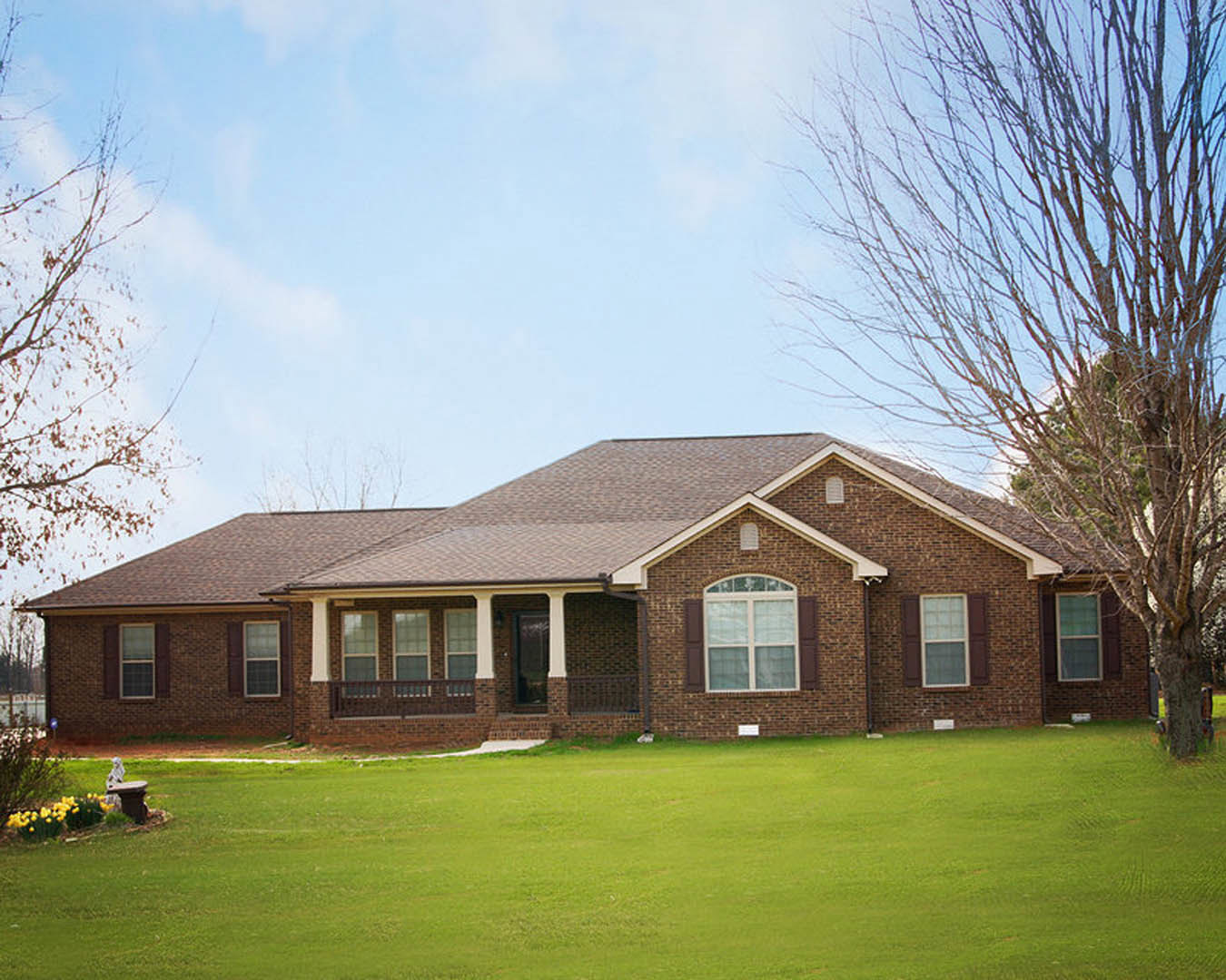 Red brick house with white-trimmed windows, covered front porch, green lawn, leafless tree, and partly cloudy sky