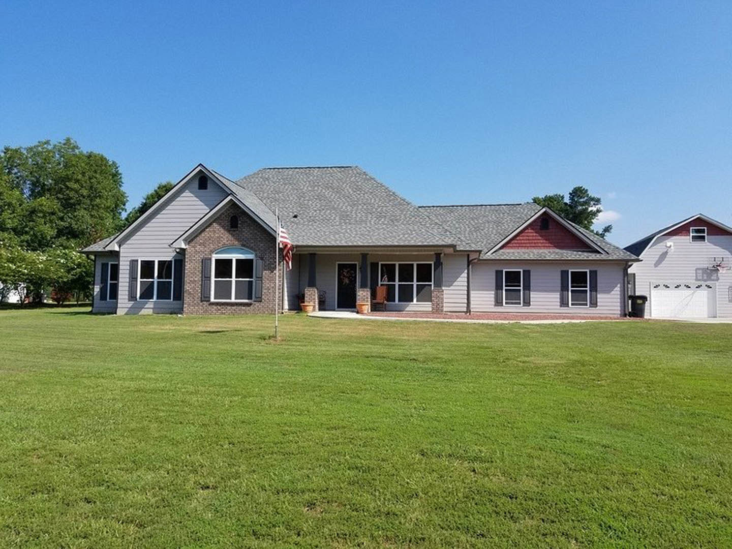 Two-story farmhouse with white siding, black front door, white-framed windows, American flag hanging near entry, manicured lawn, mature tree, blue sky in background