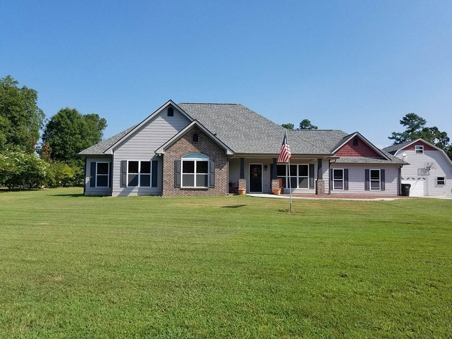 White farmhouse with black and white window frames, covered front porch, American flag on a pole, manicured lawn, mature trees, blue sky in background