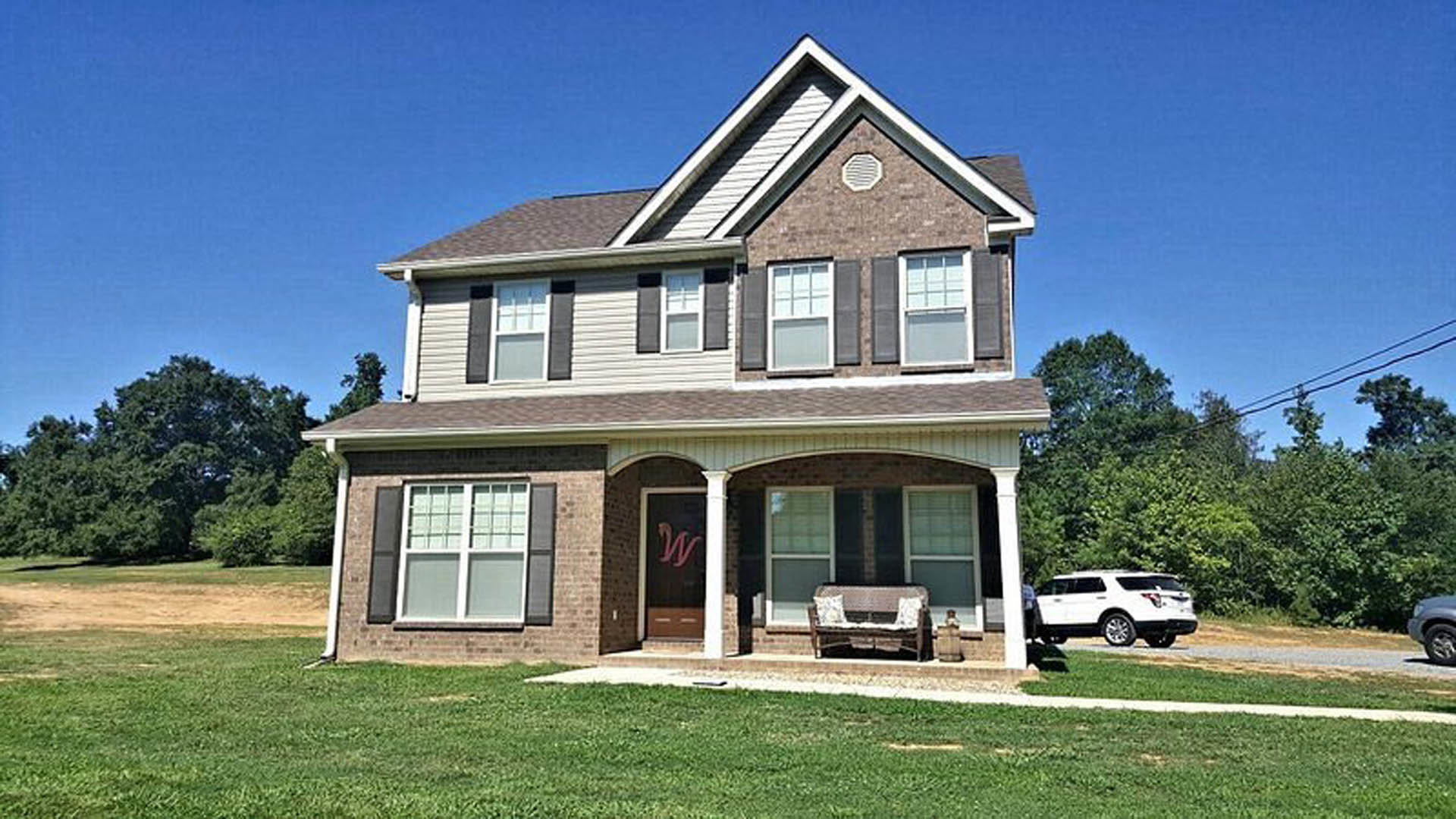 Two-story house with light siding, large windows, covered porch, green lawn, and a white car parked in driveway; trees and blue sky in background.