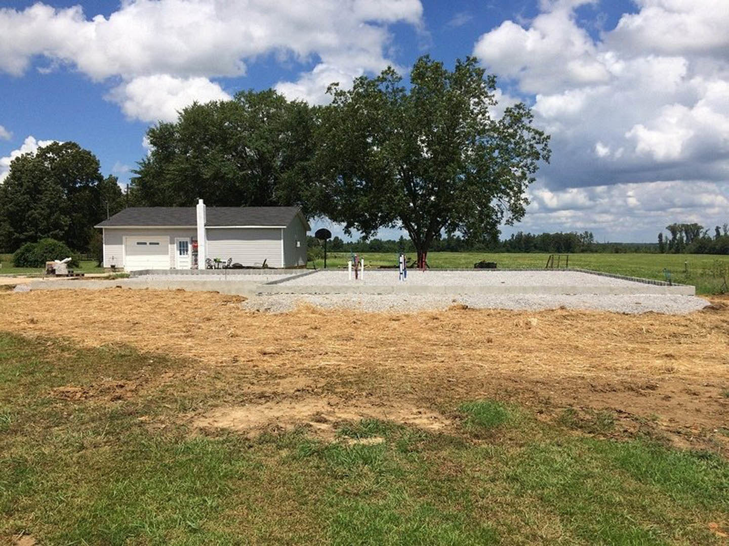 White house with prominent chimney, surrounded by dirt and grass, basketball hoop near fenced grassy field, mature trees in background under partly cloudy blue sky