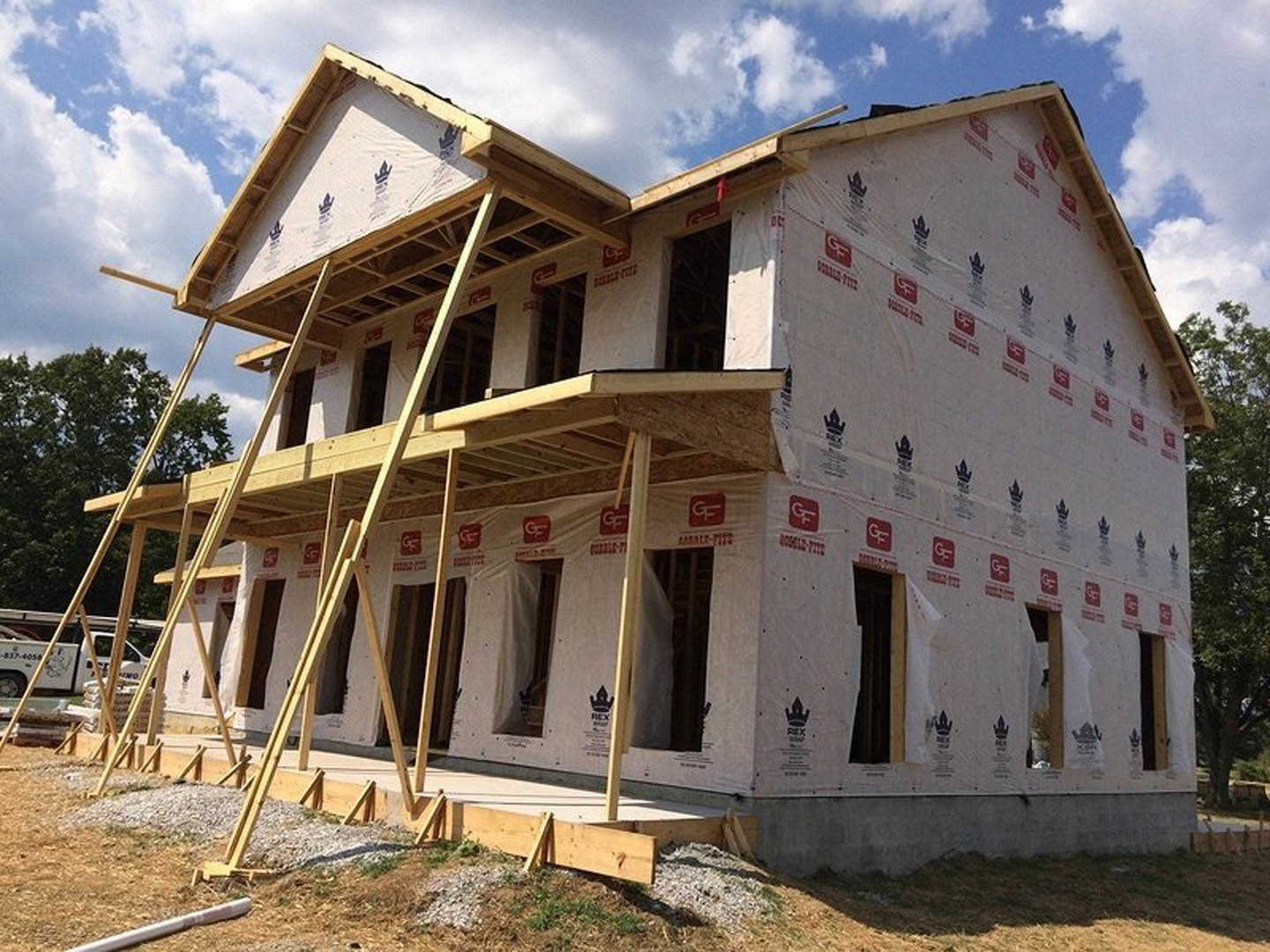 Two-story house framed with lumber and surrounded by metal scaffolding, cloudy sky overhead, trees in background, white van parked nearby.