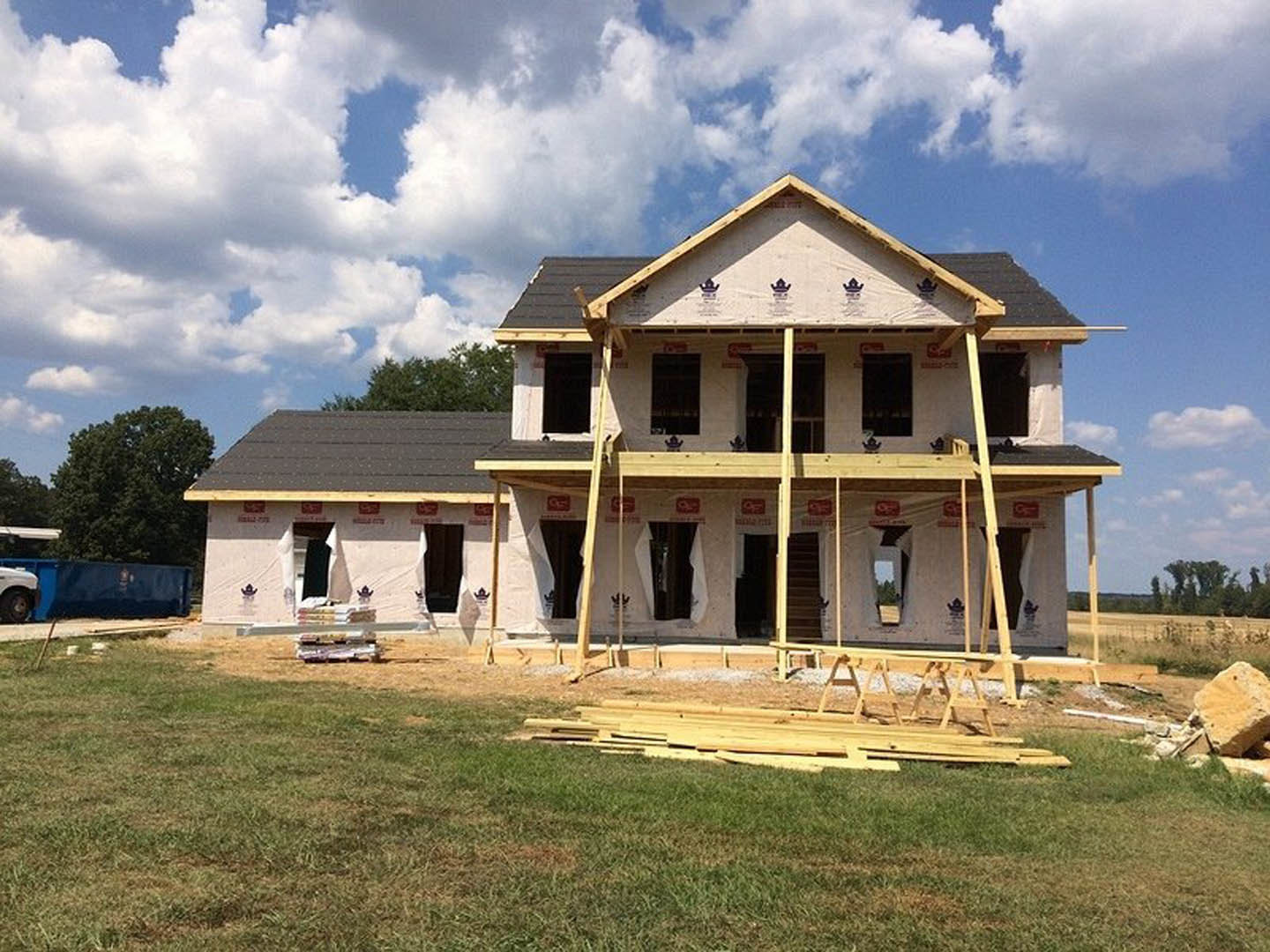 Partially built house with exposed wooden framing, scattered planks on green lawn, scaffolding along exterior, staircase visible inside, cloudy sky overhead, Grouseland mansion in