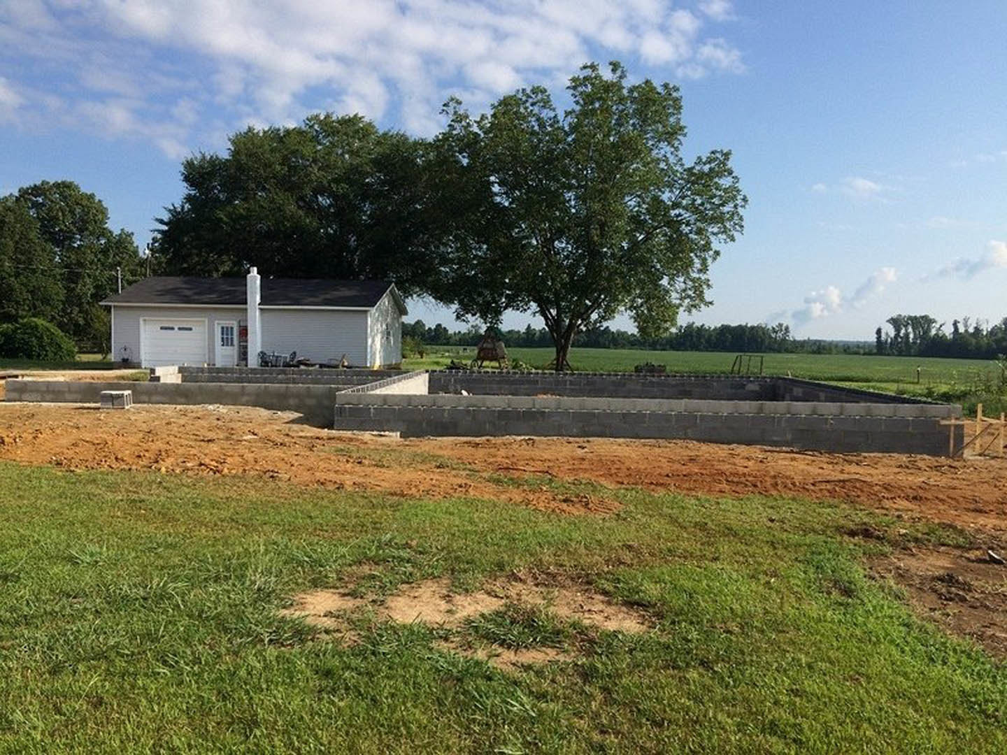 Partially built house with exposed framing, white chimney, brick steps, dirt and grass yard, mature tree in background
