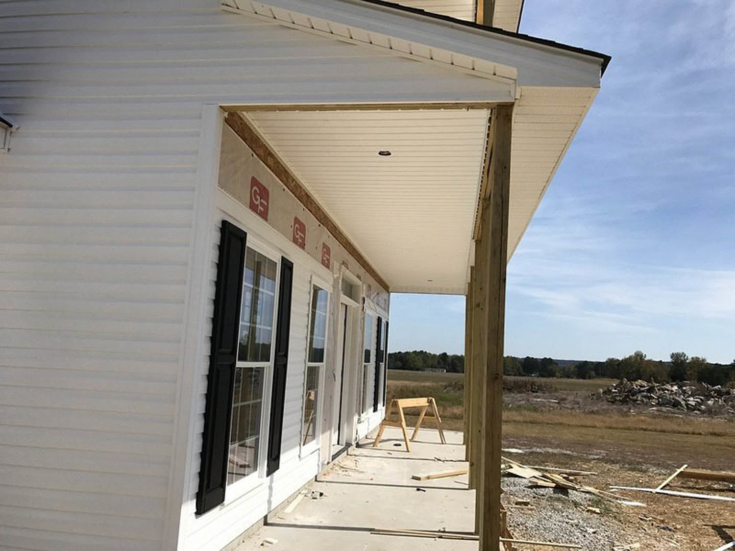 White house under construction with exposed framing, concrete porch, wooden sawhorse, pile of rocks, grassy field, and cloudy sky in background