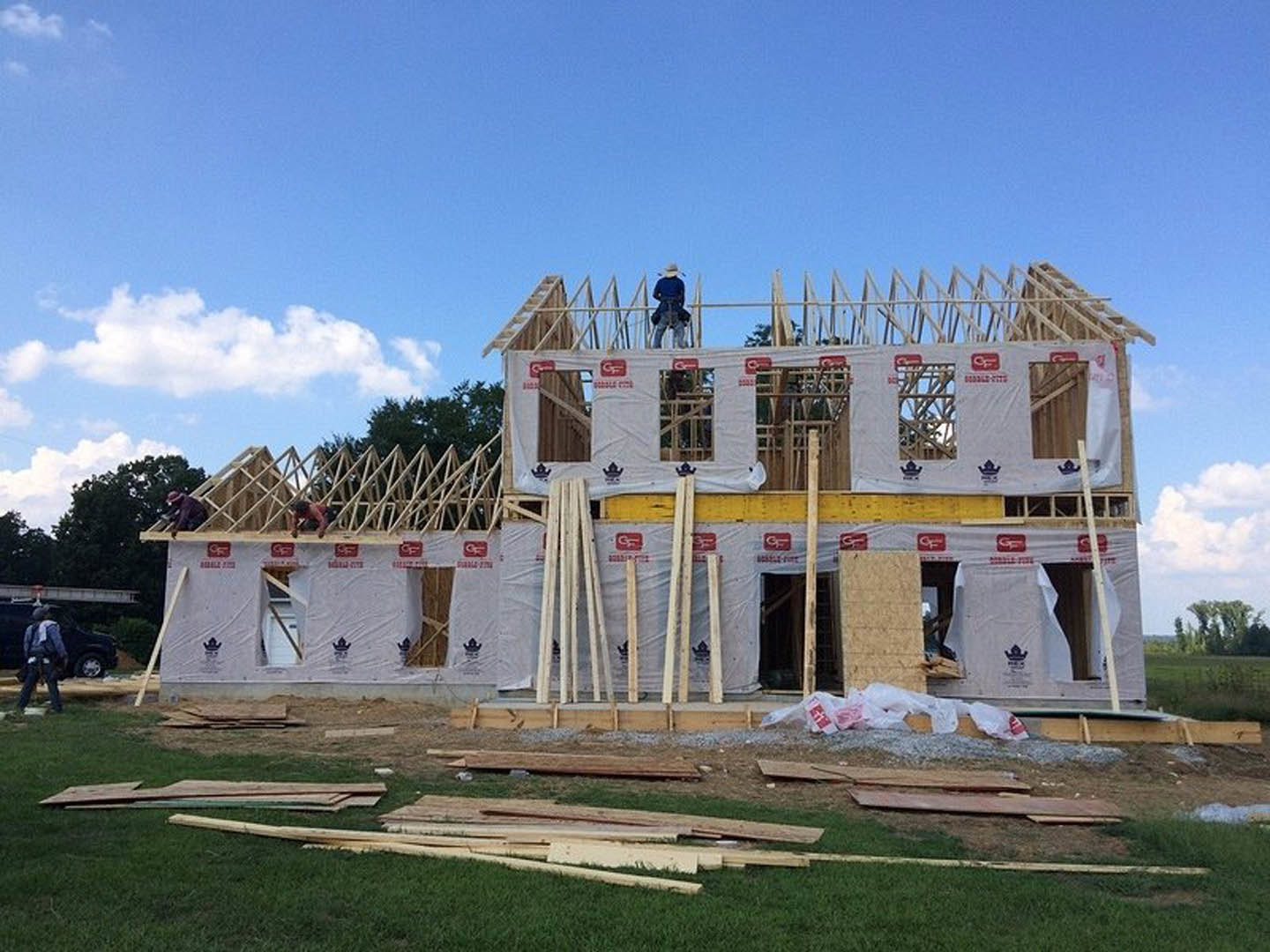 Framed wooden house under construction with exposed beams, surrounded by grass and trees, cloudy sky overhead