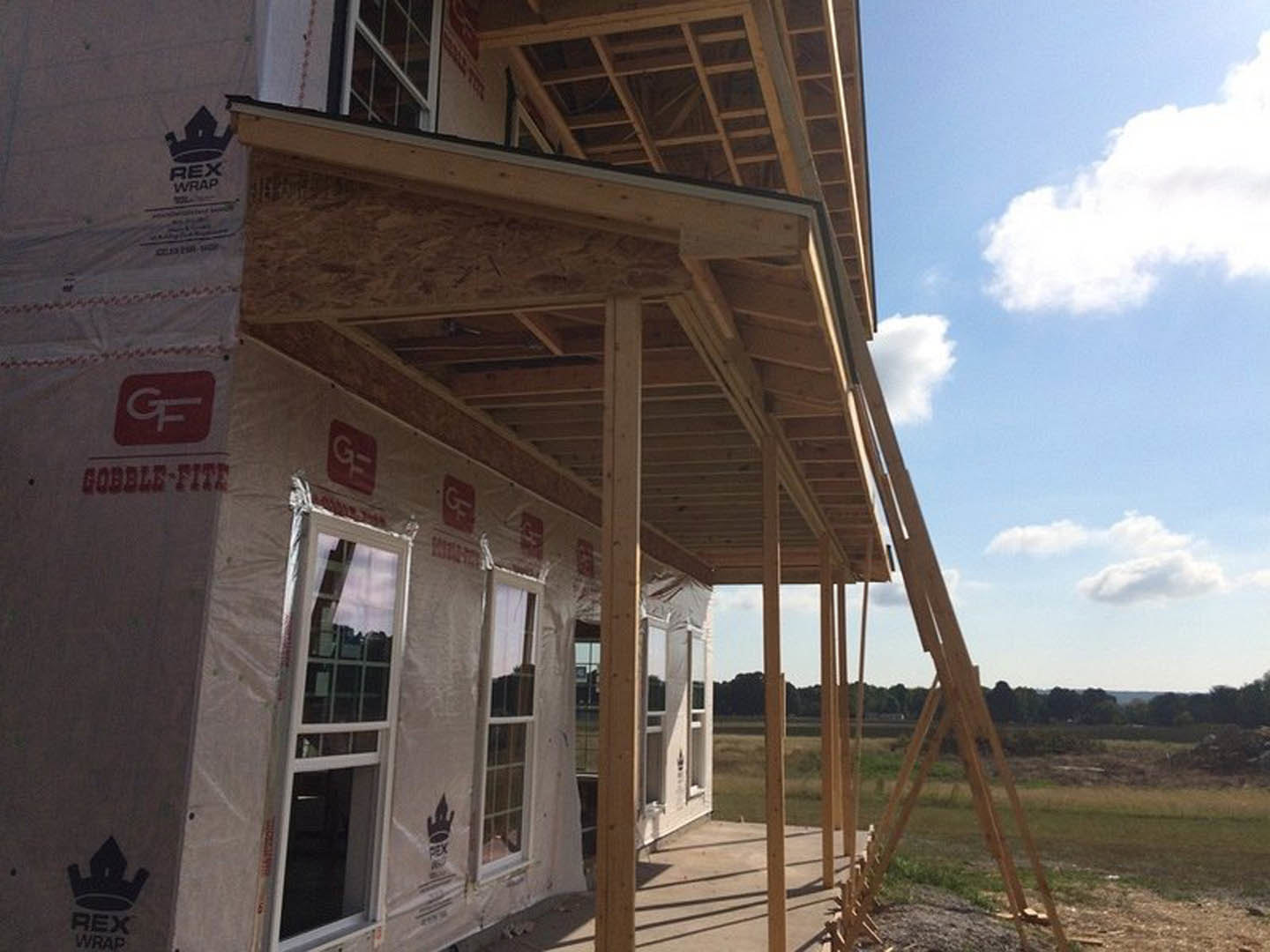 Two-story house under construction with metal scaffolding, glass windows installed, red and white construction sign on exterior wall, grassy ground, partly cloudy sky overhead