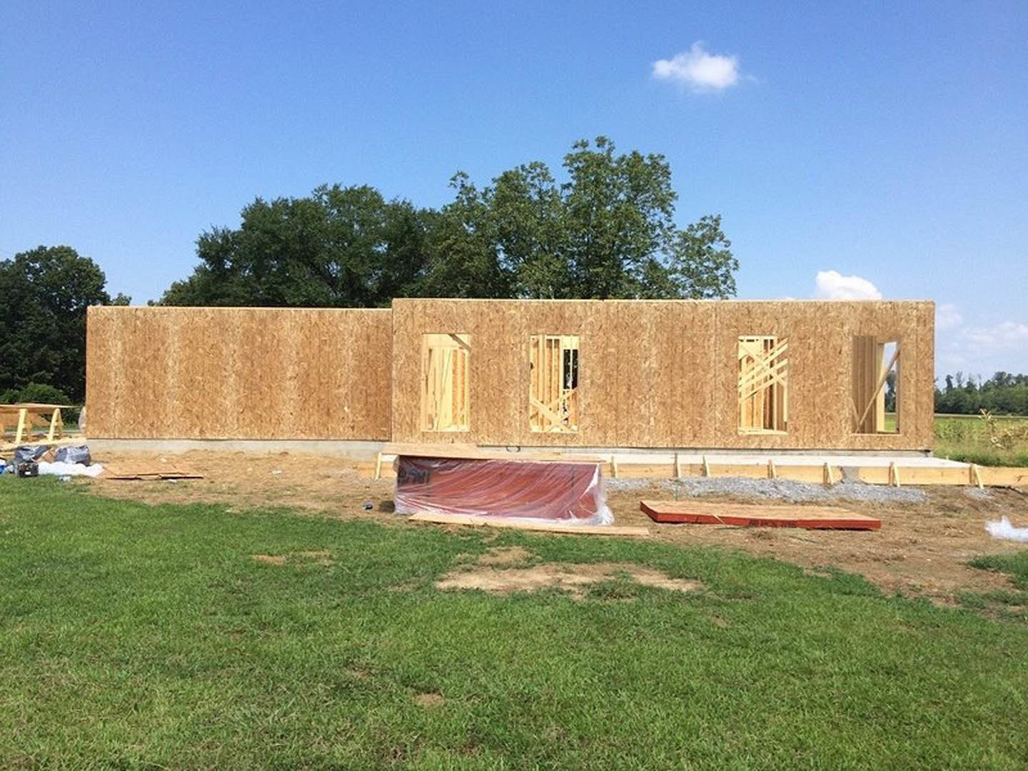 Partially built house with exposed framing, red sheathing panels, large windows, surrounded by green grass and mature trees under a cloudy sky