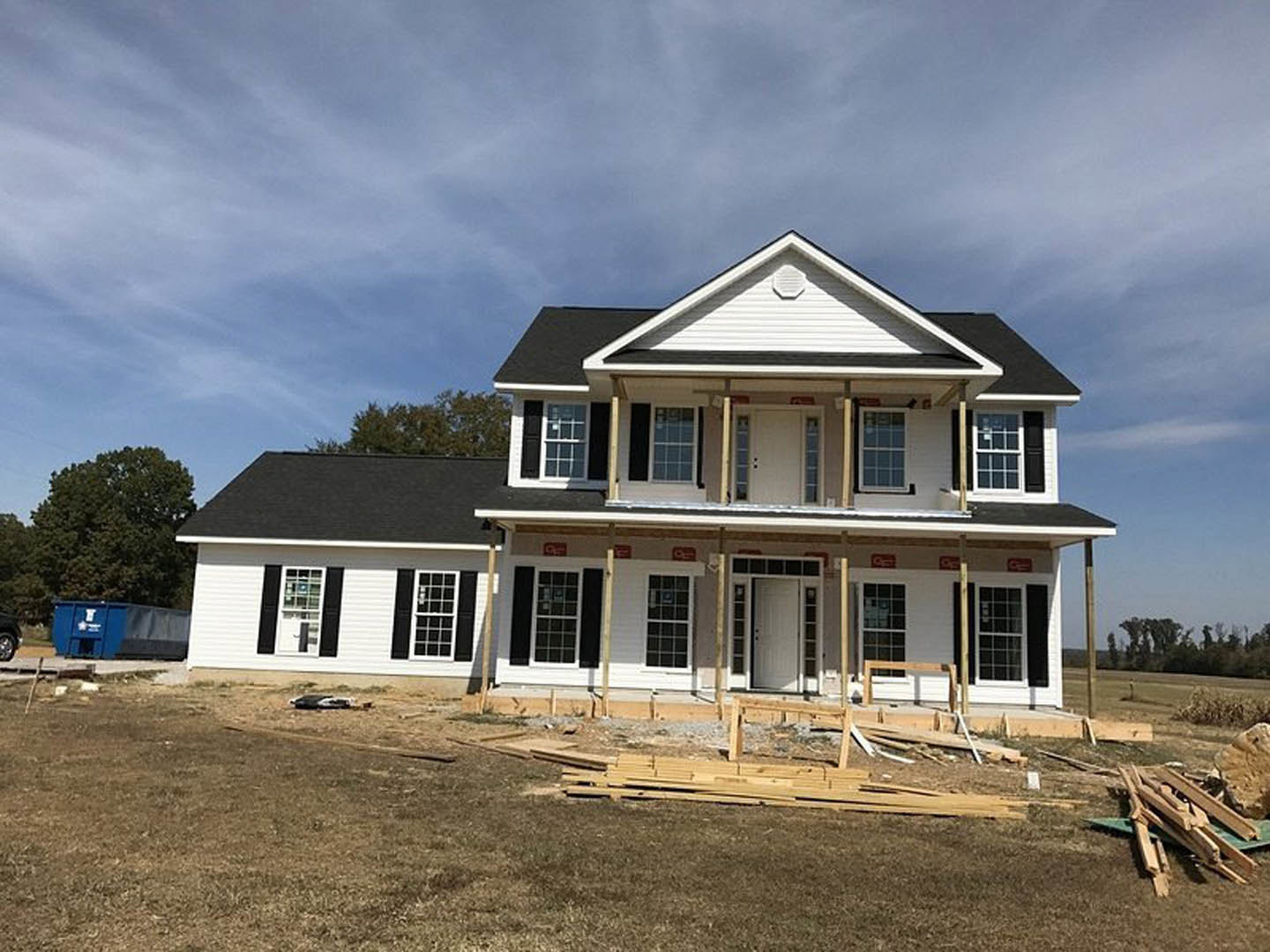 Partially built house with exposed wooden framing, white window frame, pile of lumber on dirt, blue dumpster with white lettering, green-leafed tree nearby, open field and cloudy