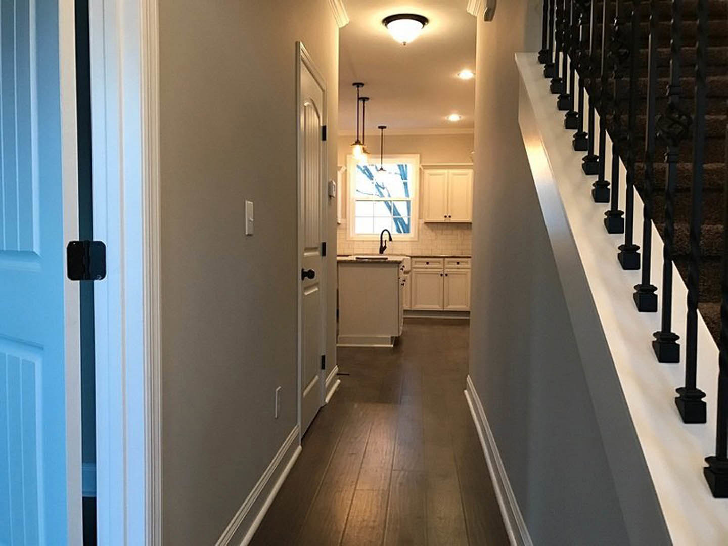 Hallway with wood flooring, white walls, black metal stair railings, a white cabinet, ceiling light fixture, and a closed door at the end