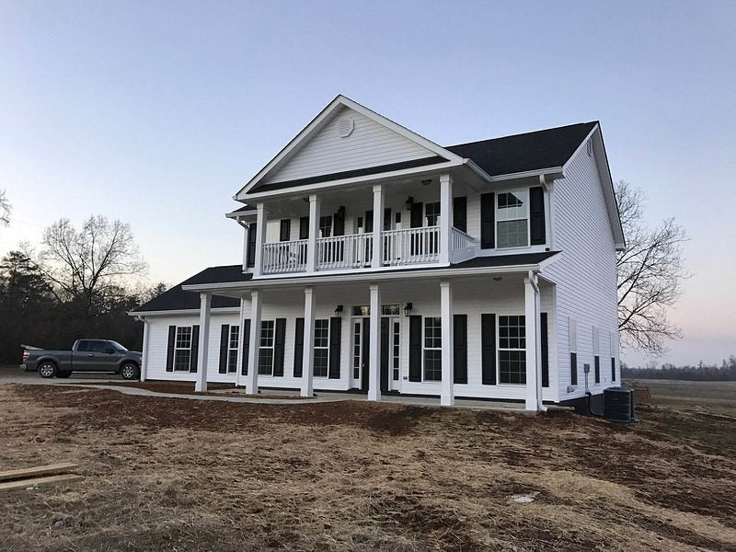 White house with black shutters, white porch railing, brown dirt patch in front, parked truck, trees and sky in background