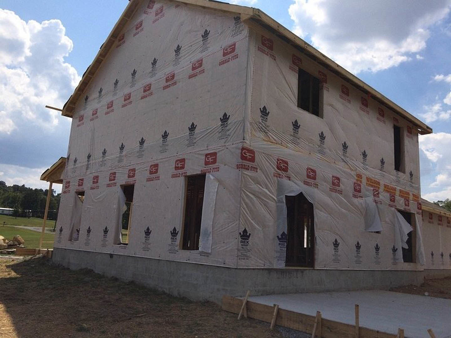 Modern home exterior under construction with plastic sheeting covering windows and entry, black-framed window, exposed wooden framing, and unfinished ground.