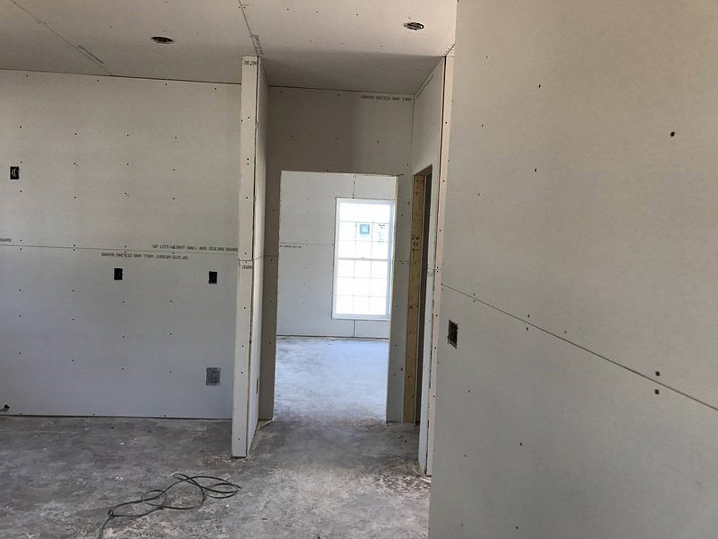 White-walled room with open door, window with white panel, exposed black wire on floor, holes in wall and ceiling, unfinished plaster surfaces