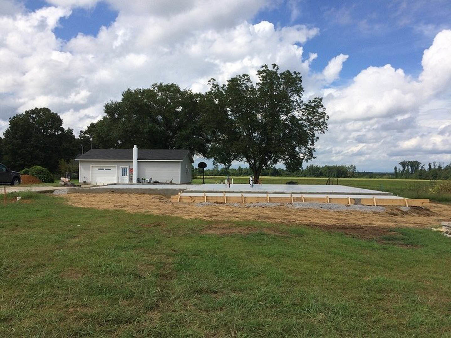 White house with chimney, concrete slab and foundation in front, green lawn, trees in background, cloudy sky overhead