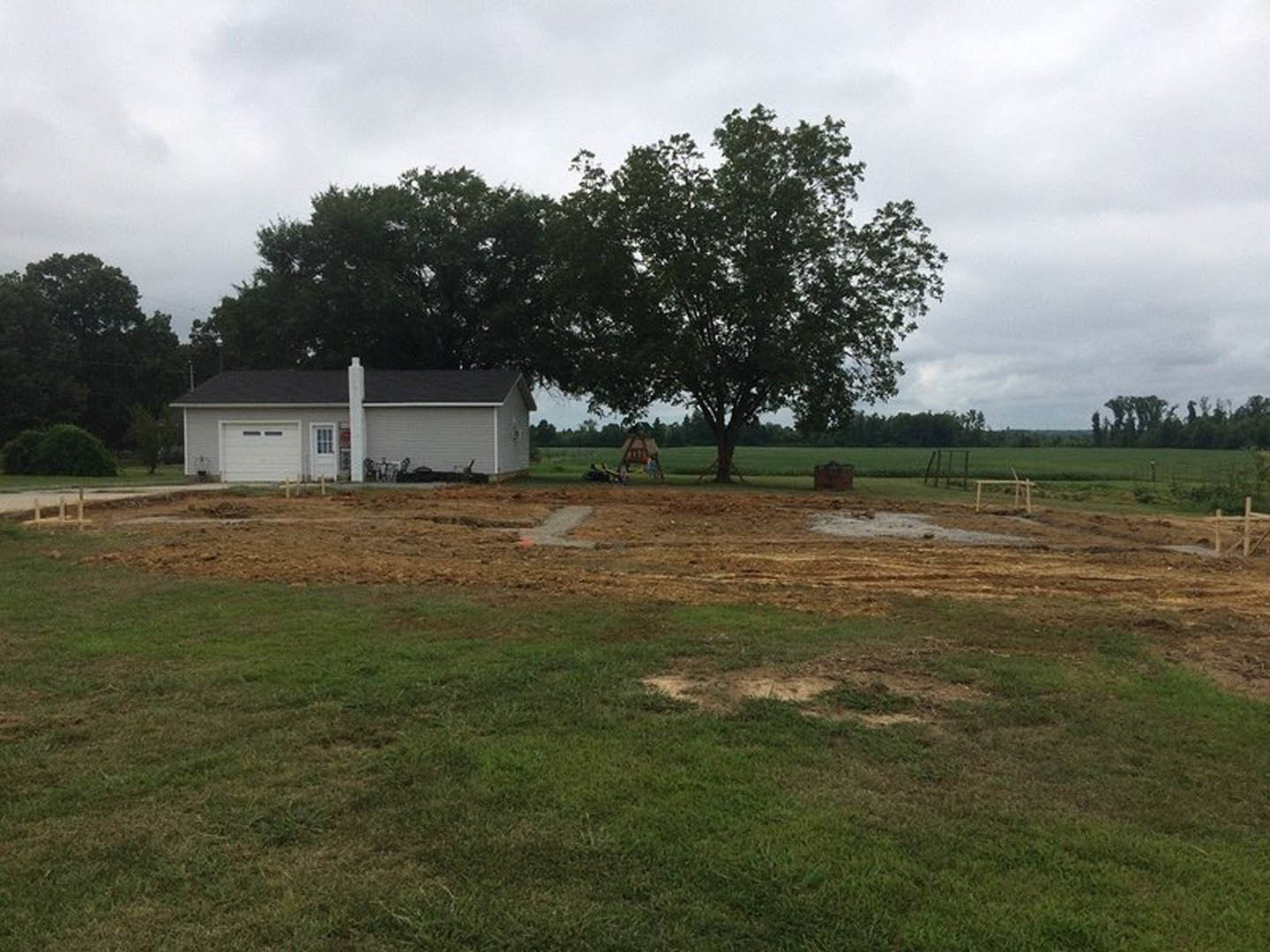 Two-story house with brick chimney, surrounded by grass and a large patch of bare dirt, mature tree nearby, cloudy sky overhead