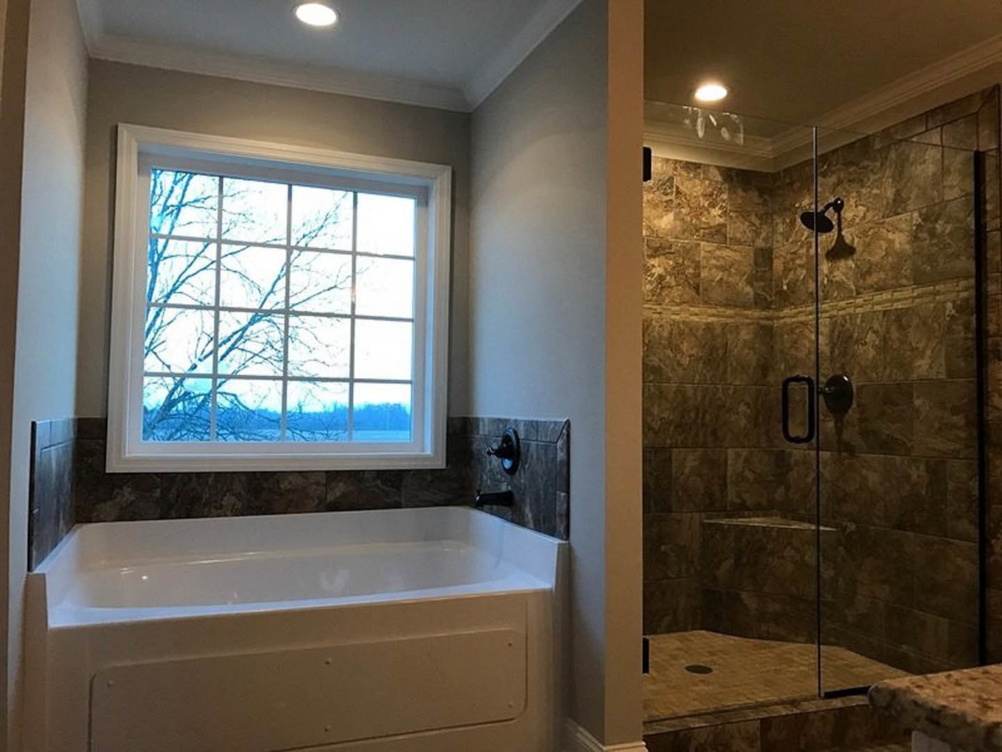 Freestanding white bathtub beneath a large window with tree view, glass-enclosed shower with stone tile, white vanity with glass countertop, chrome fixtures, and light tile
