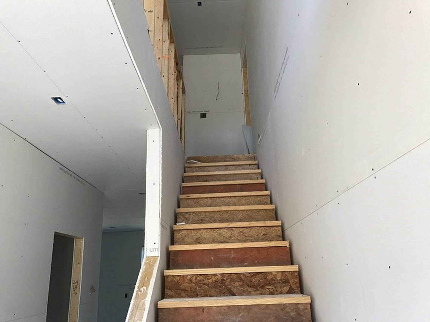 Wooden staircase with light-toned treads and white plaster walls, black handrail, and a white curtain partially visible in the background.