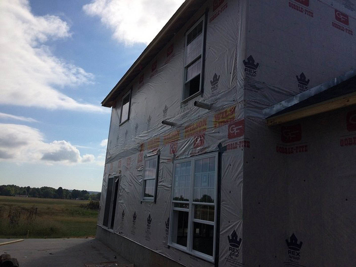 Modern home exterior under construction with plastic sheeting covering windows, black window frames, grassy yard, and distant trees under cloudy sky