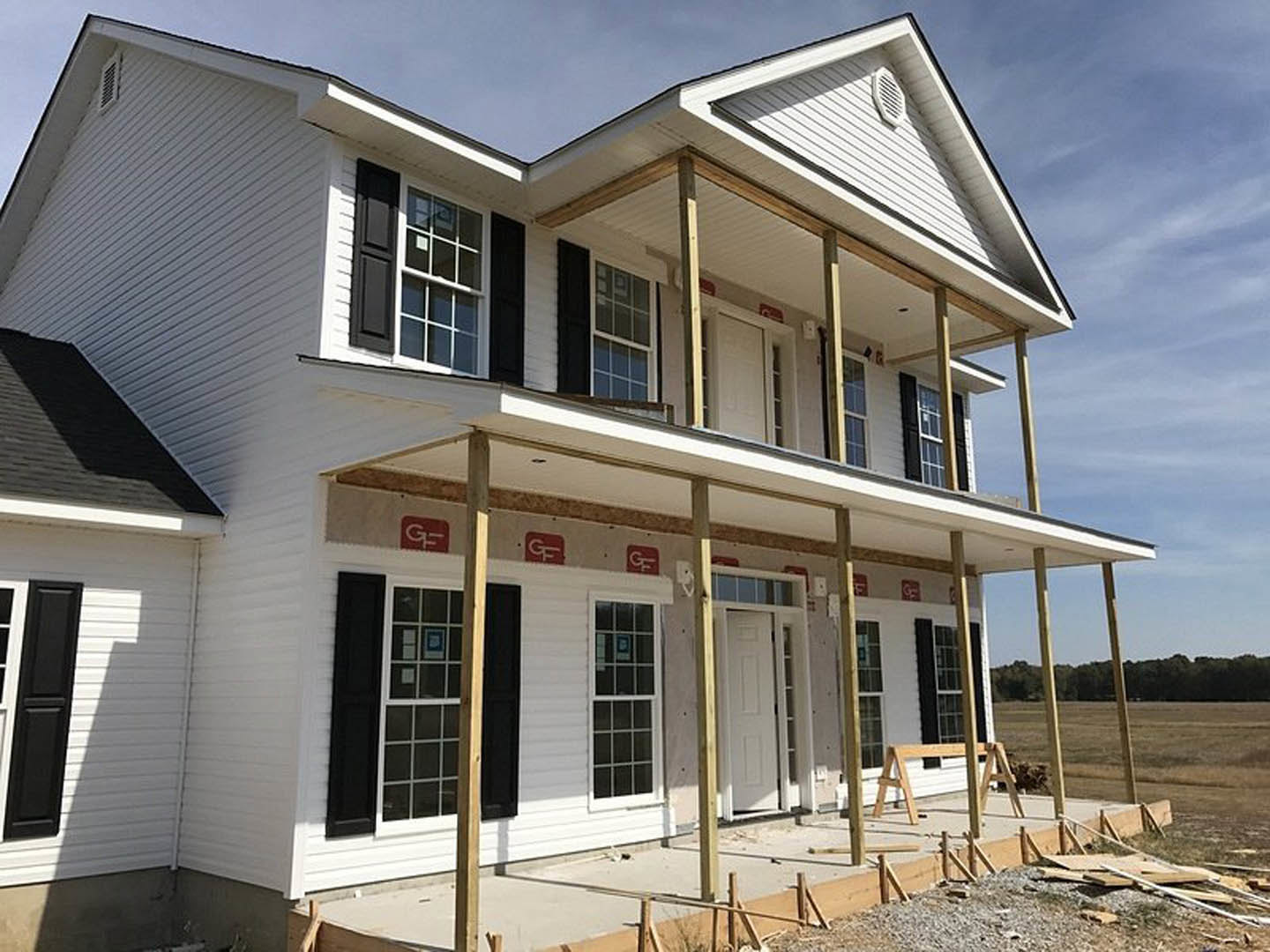 White siding house under construction with exposed porch pillars, multi-pane windows, black shutters, glass-paneled white door, and wooden bench on sidewalk