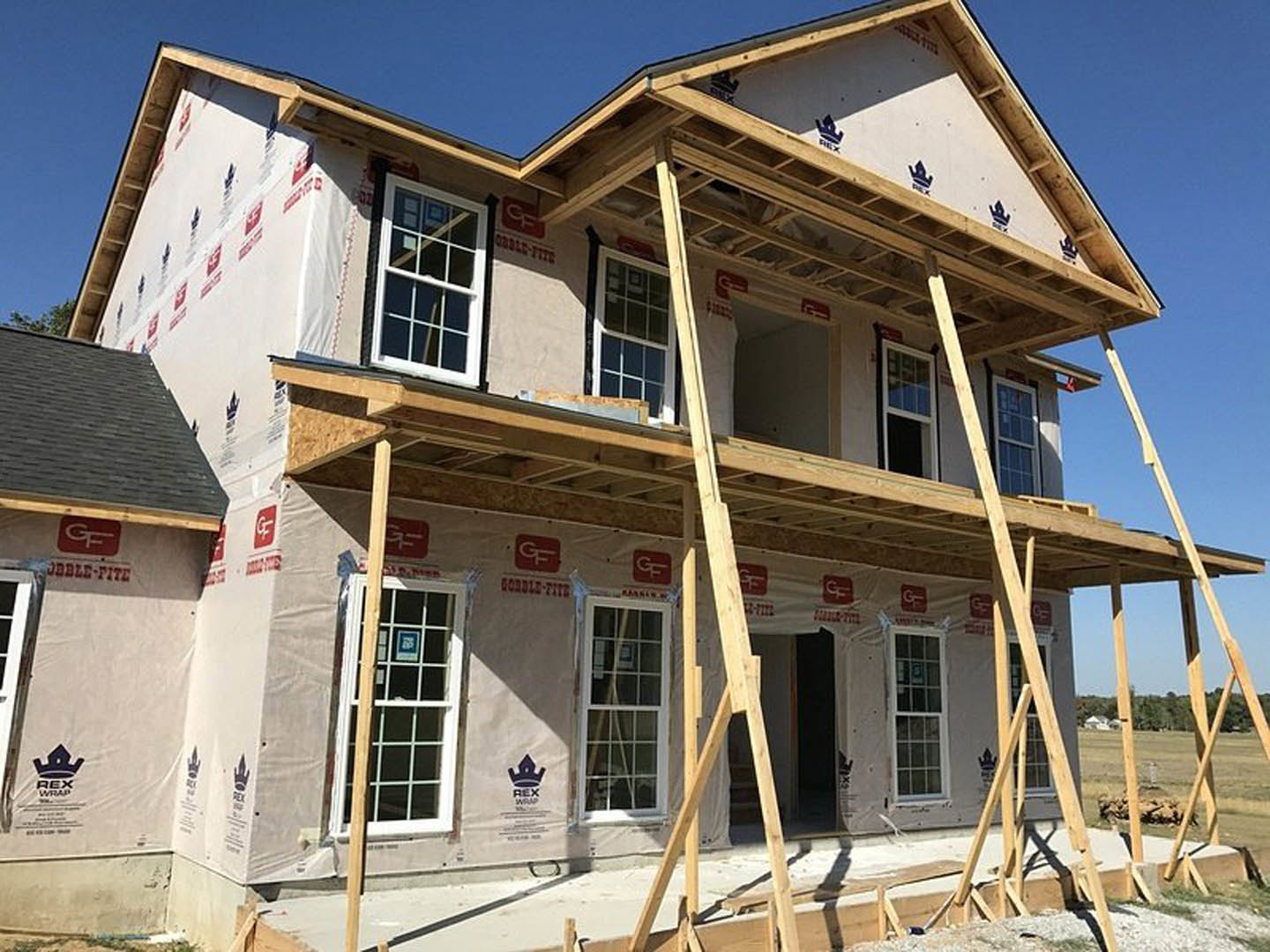 Two-story house under construction with wood scaffolding, white-framed window displaying a company logo sign, exposed siding and lumber, blue sky in background