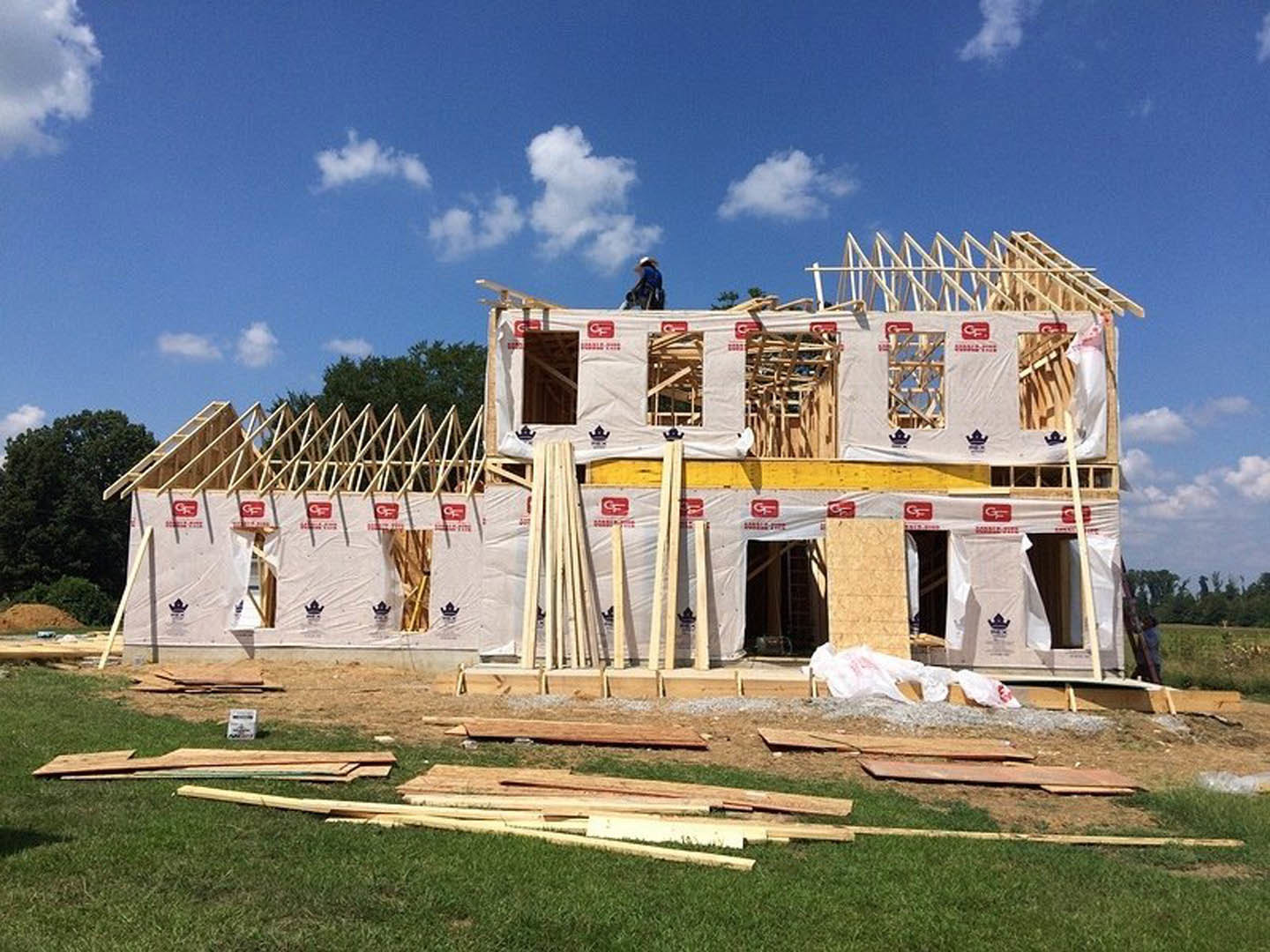 Framed house under construction with exposed lumber, unfinished roof, grassy lot, and scattered wood piles.