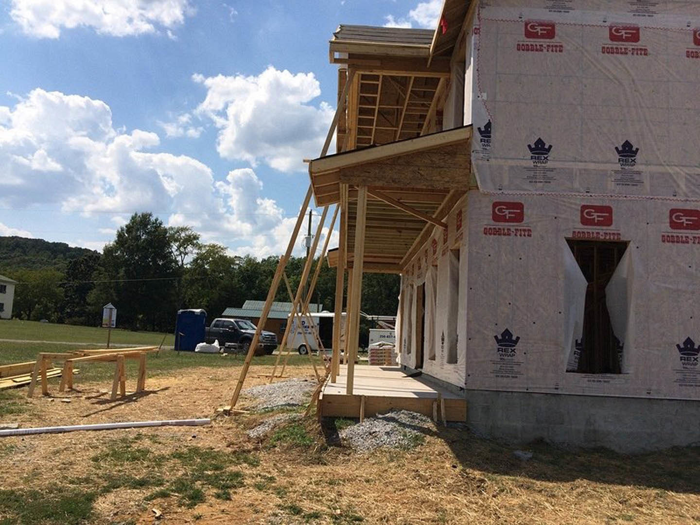 Partially built house with exposed wooden framing, wooden ladder leaning against structure, blue portable toilet on snowy ground, red and white sign with letter "F", parked car
