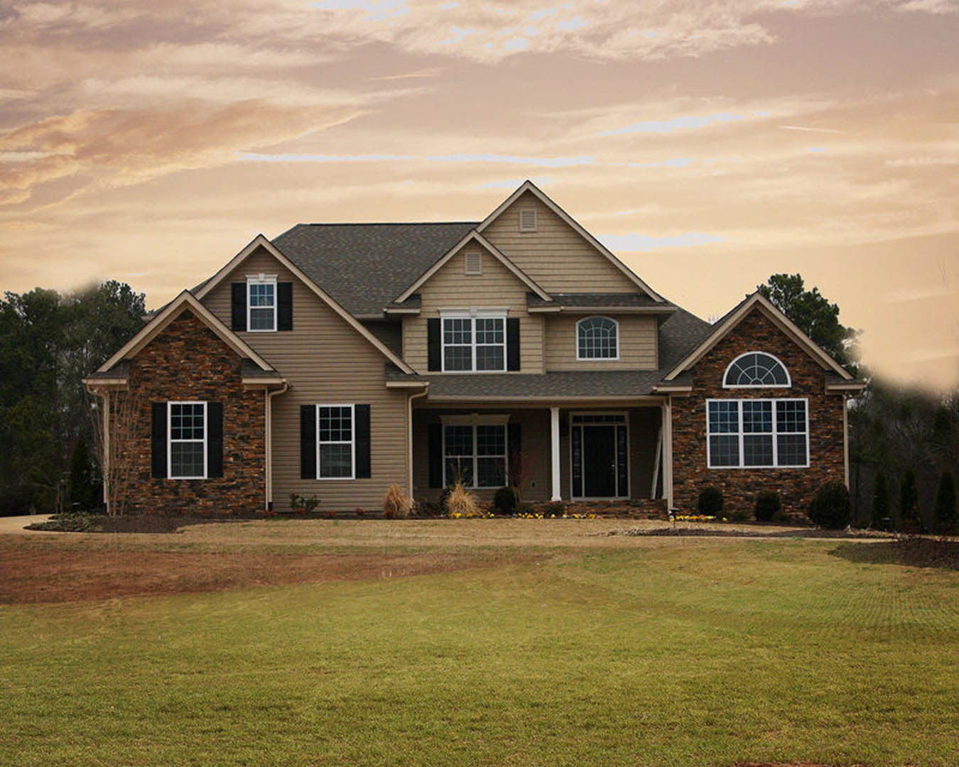 Two-story farmhouse with white-framed windows, gray siding, and a spacious green lawn bordered by trees under a partly cloudy sky
