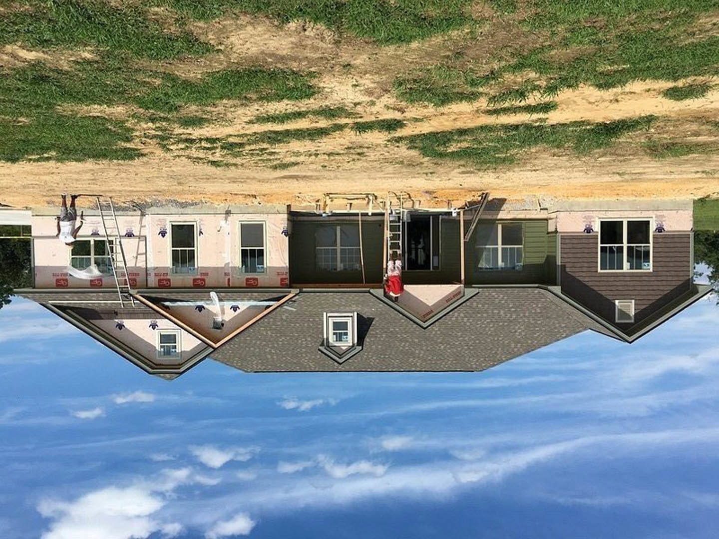 Partially built house with exposed framing, white window frame, man on ladder working on roof, dirt road bordered by grass, clear blue sky overhead