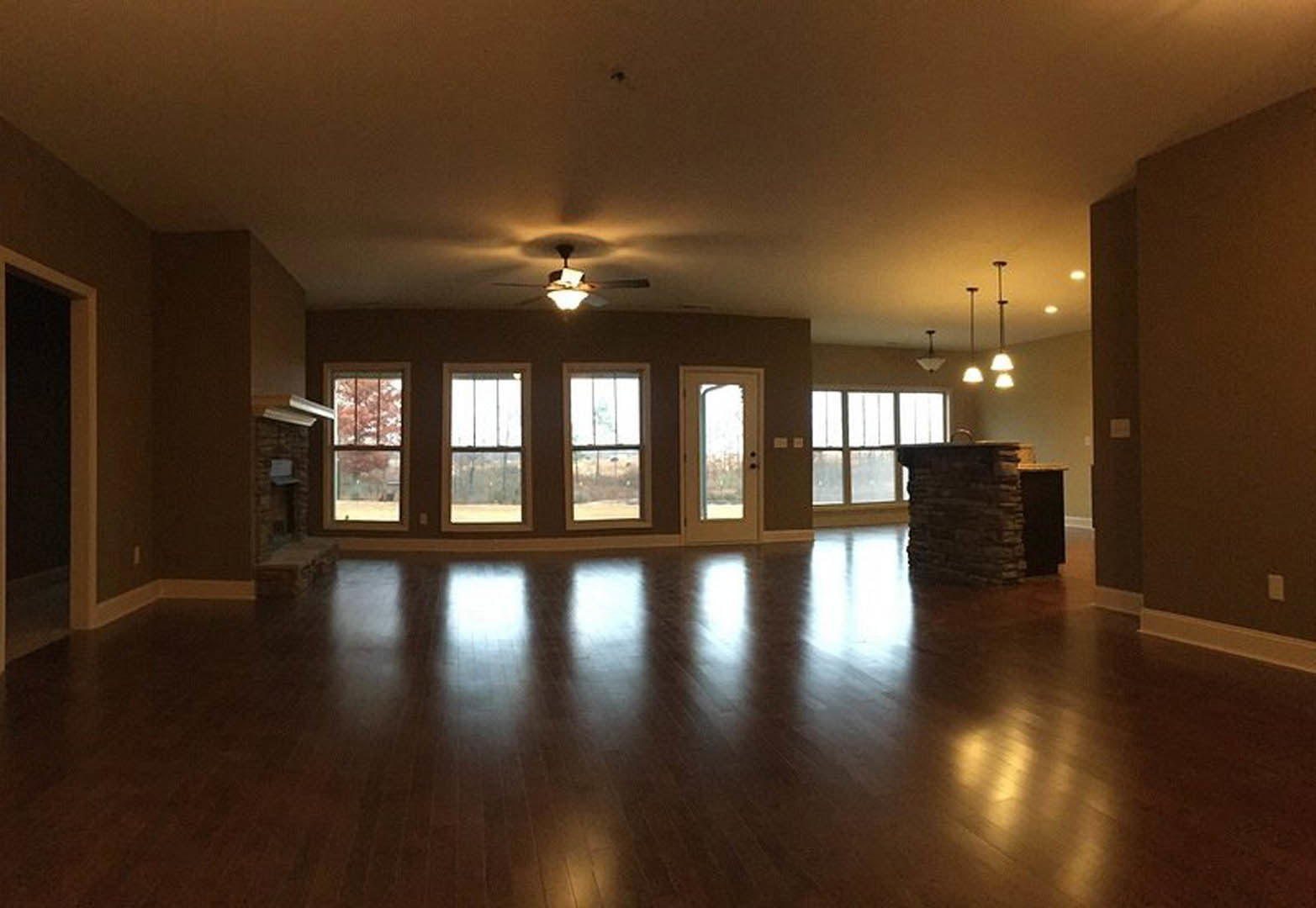 Spacious living room featuring a stone fireplace, hardwood floors with light reflections, ceiling fan, multiple windows, and a door with glass inset.