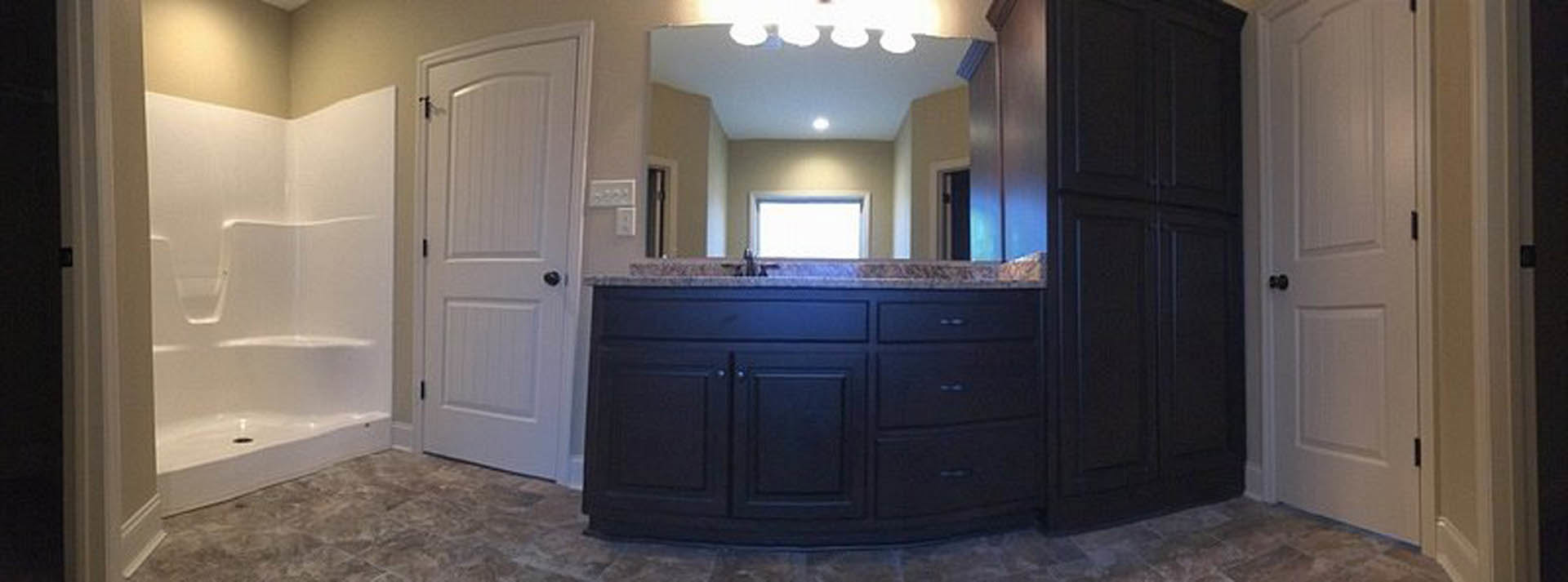 White bathroom featuring a large wall mirror above a black countertop, white cabinetry with black knobs, grey surface accents, and a white shower with built-in shelf.