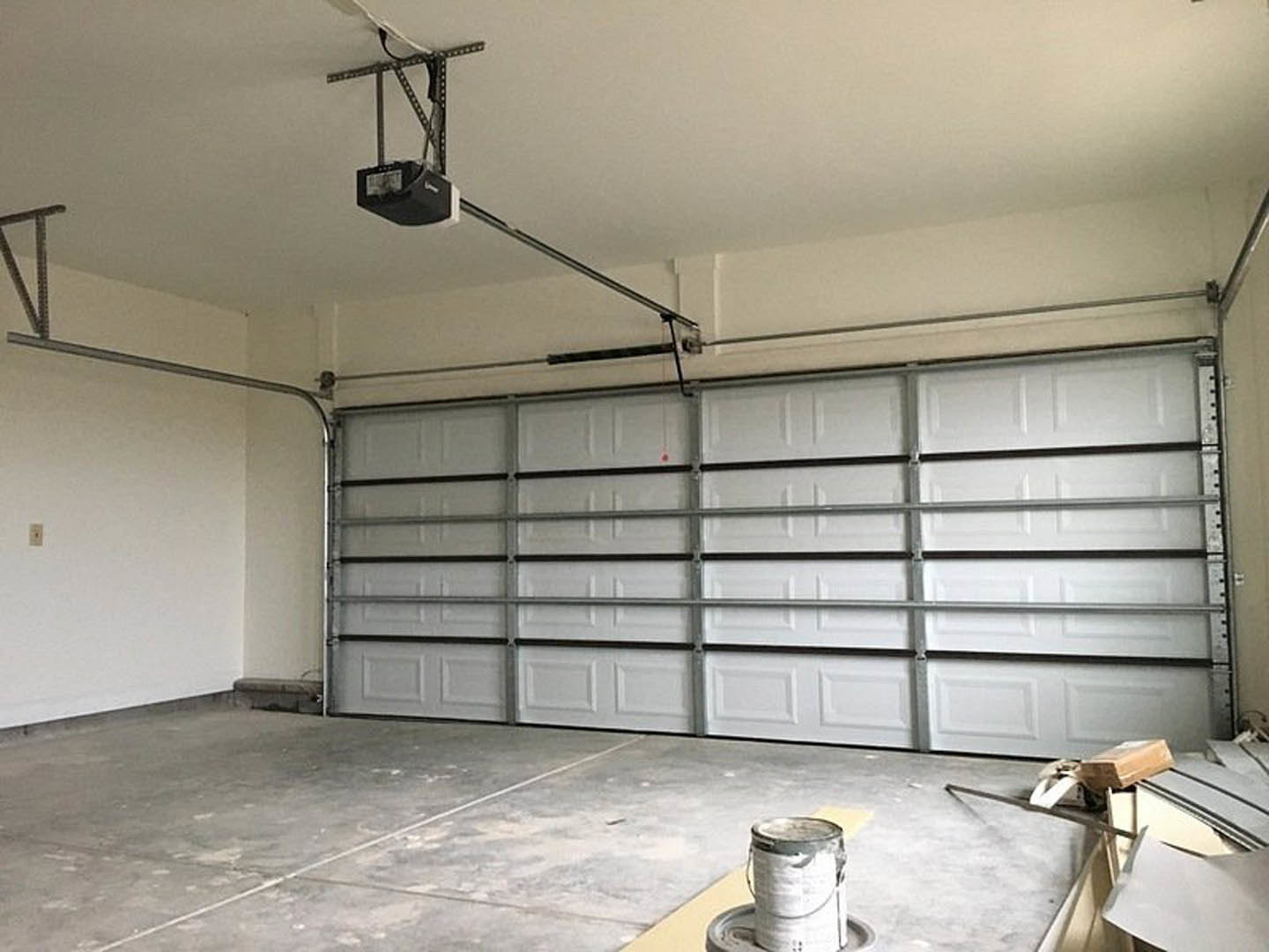 Garage interior with smooth white plaster walls, concrete floor, metal-framed garage door, ceiling fan, metal shelving holding a black box, wooden block on workbench, and paint can