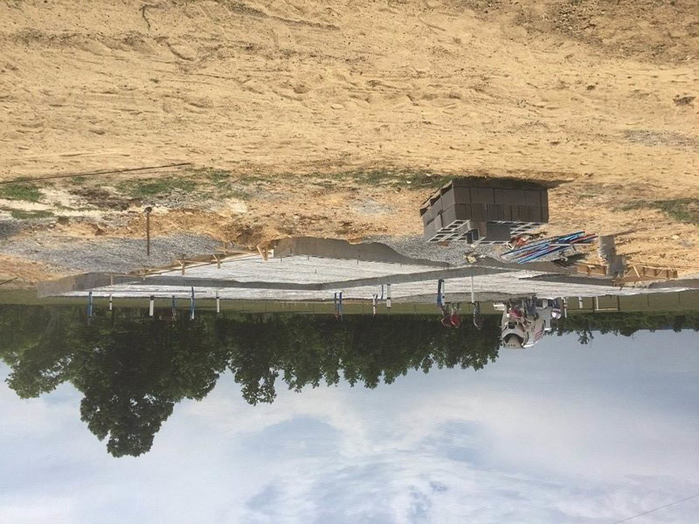 Grey brick house under construction beside a dirt road, group of people standing on sidewalk, row of trees in background, dirt field with stacked pallets