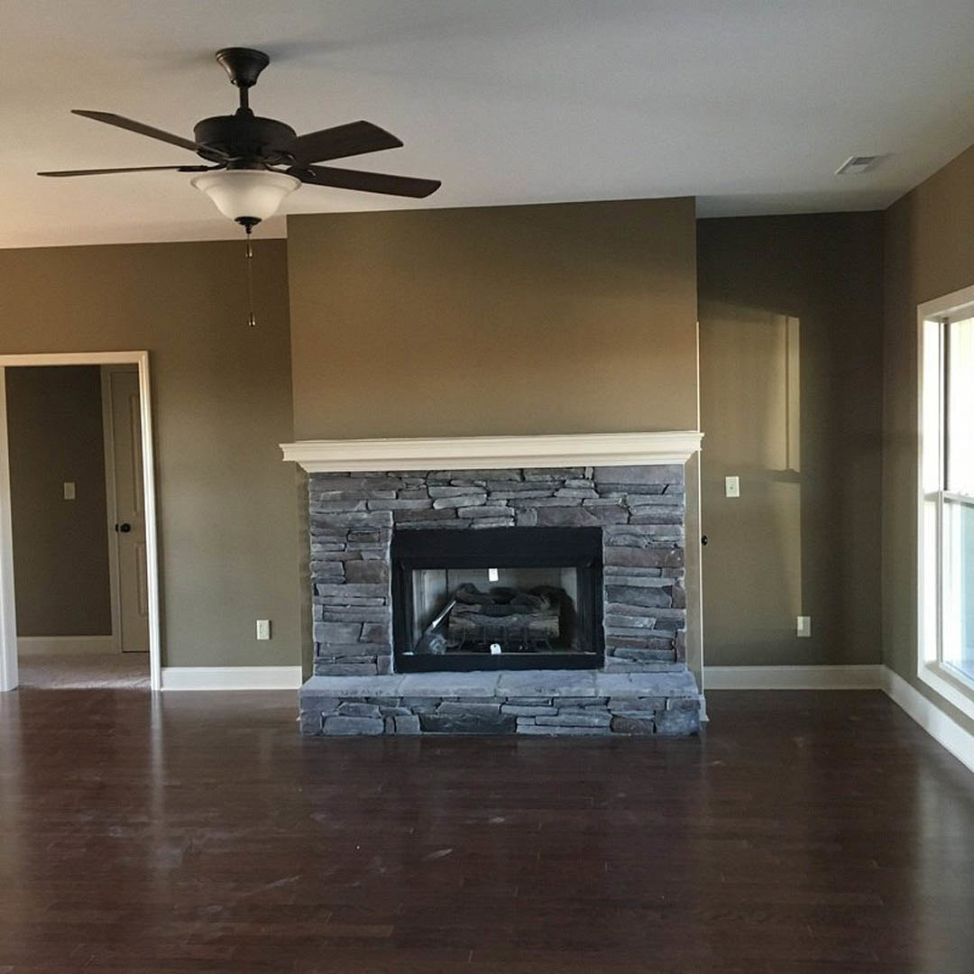 Living room with wood flooring, stone accent wall, glass-front fireplace filled with wood logs, ceiling fan with light fixture, and white door featuring a black handle