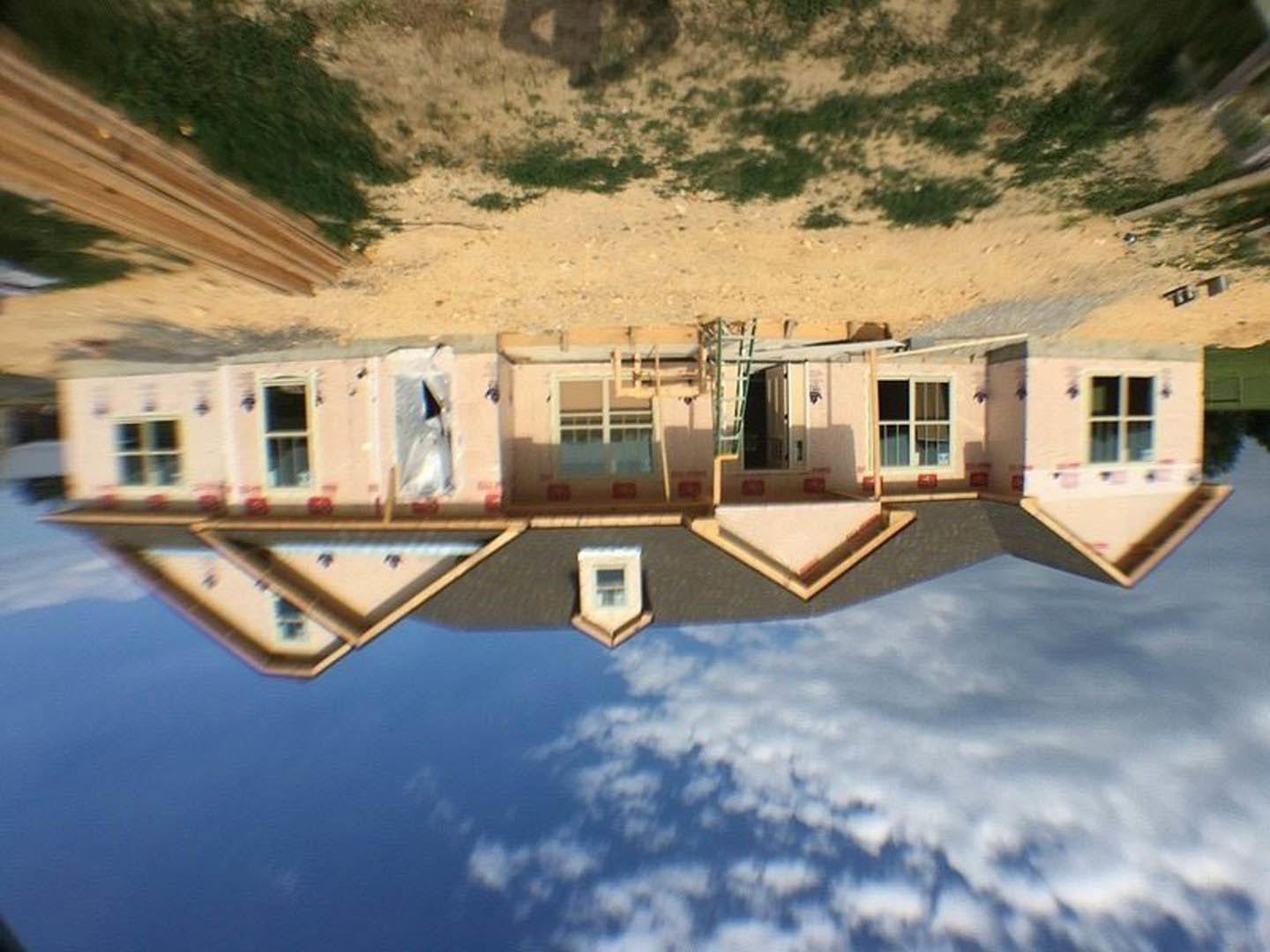 Framed custom home under construction with exposed wood, large windows reflecting blue sky and clouds, mountain landscape in background