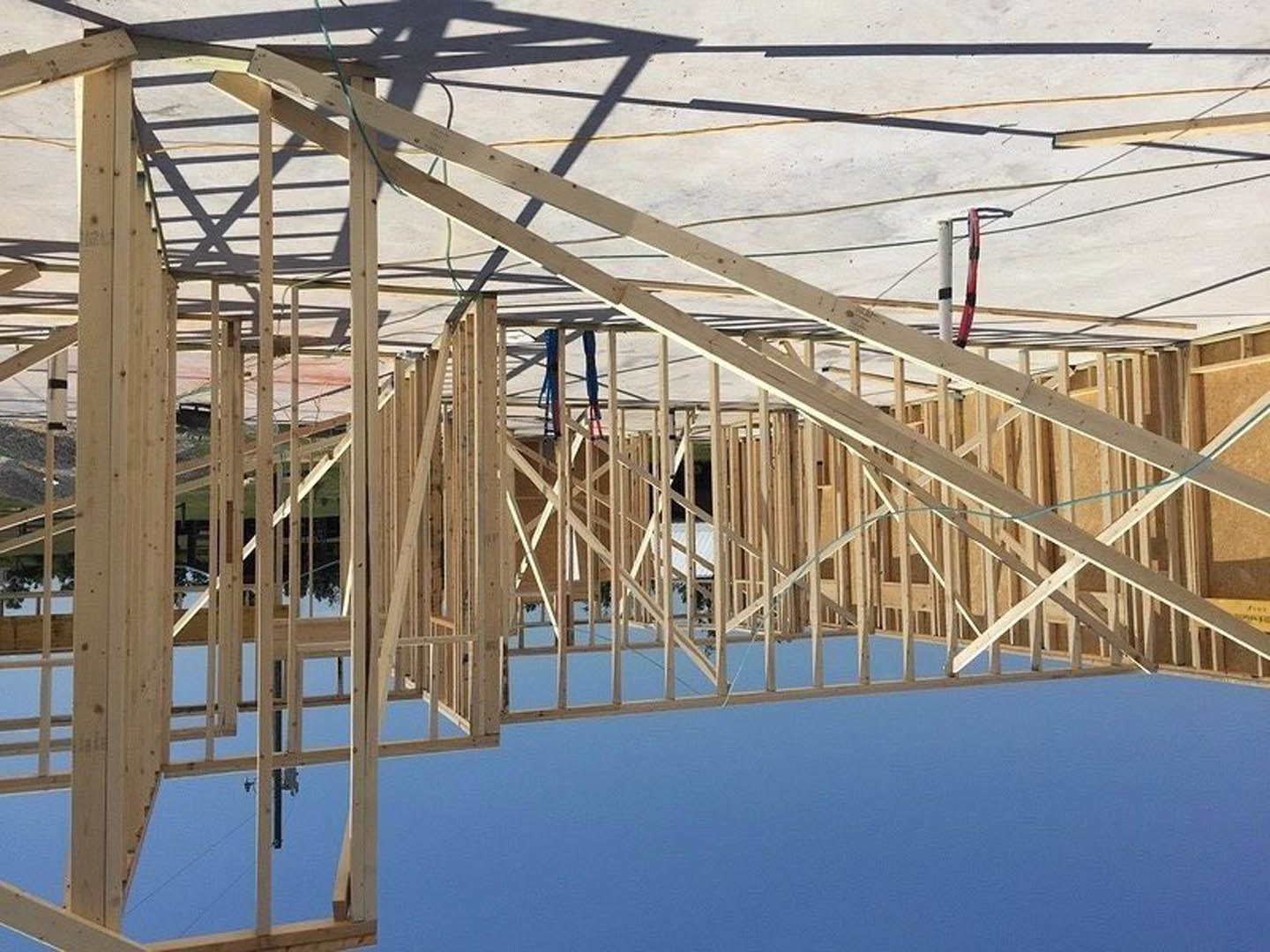 Wooden house frame under construction with exposed beams, concrete foundation, scattered planks, red and black pole, and clear blue sky in the background.