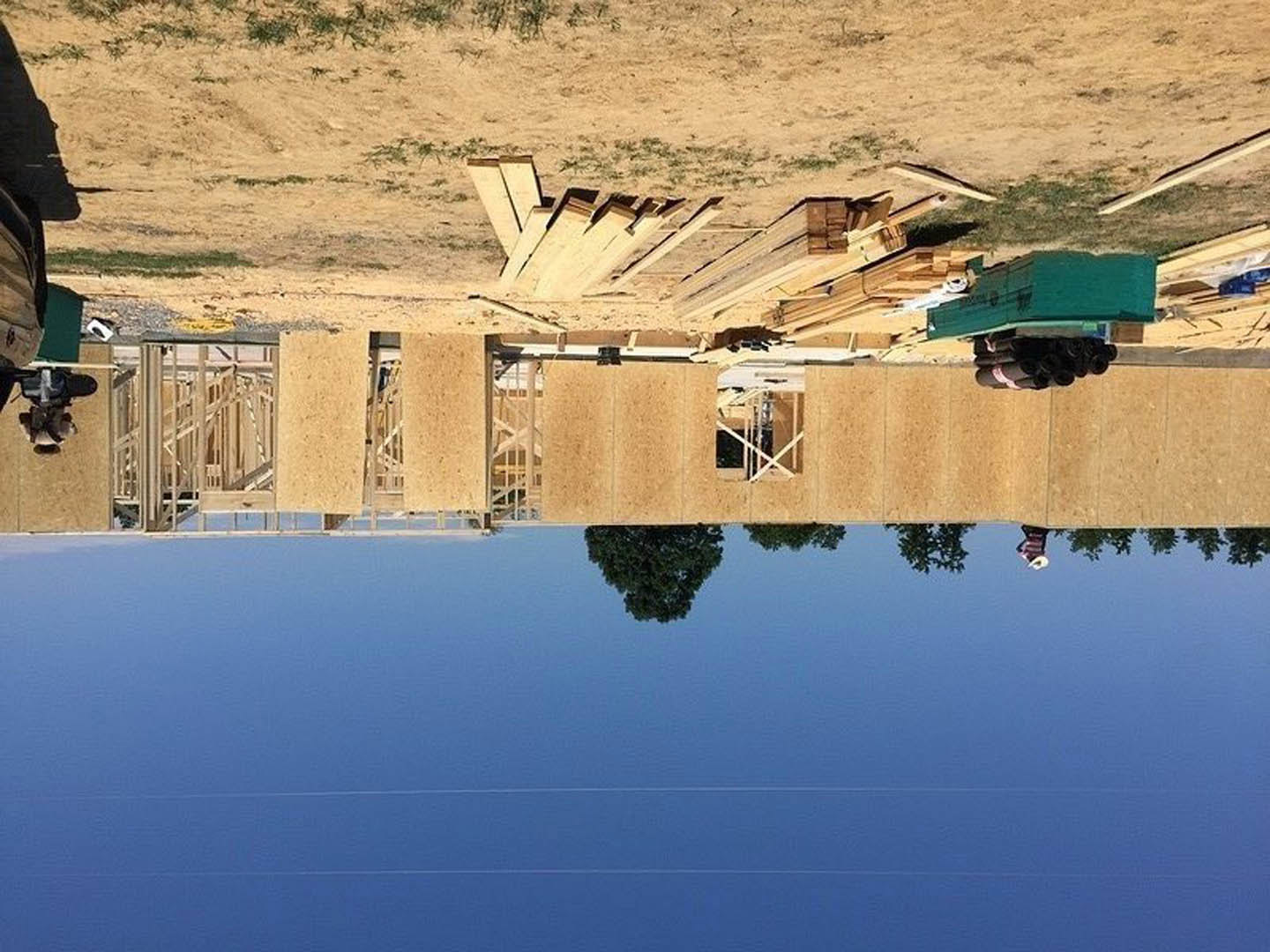 Wooden lakeside home with natural wood siding, people standing near the exterior, tree reflected in calm water, mountain backdrop, lush greenery, clear blue sky