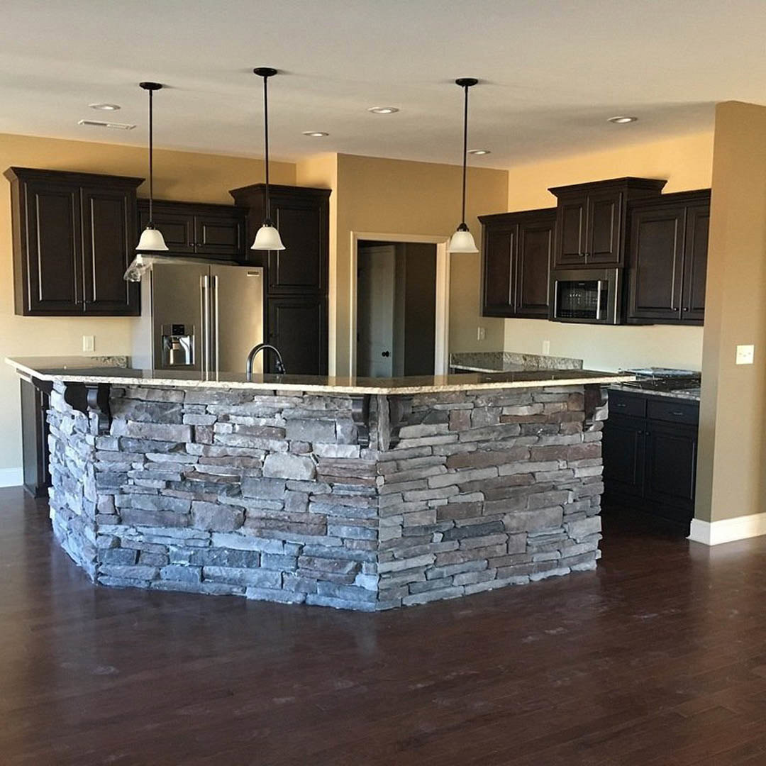 Kitchen featuring a granite island, stone accent wall, stainless steel refrigerator, glass-door microwave, dark metal support pole, white cabinetry, and light wood flooring