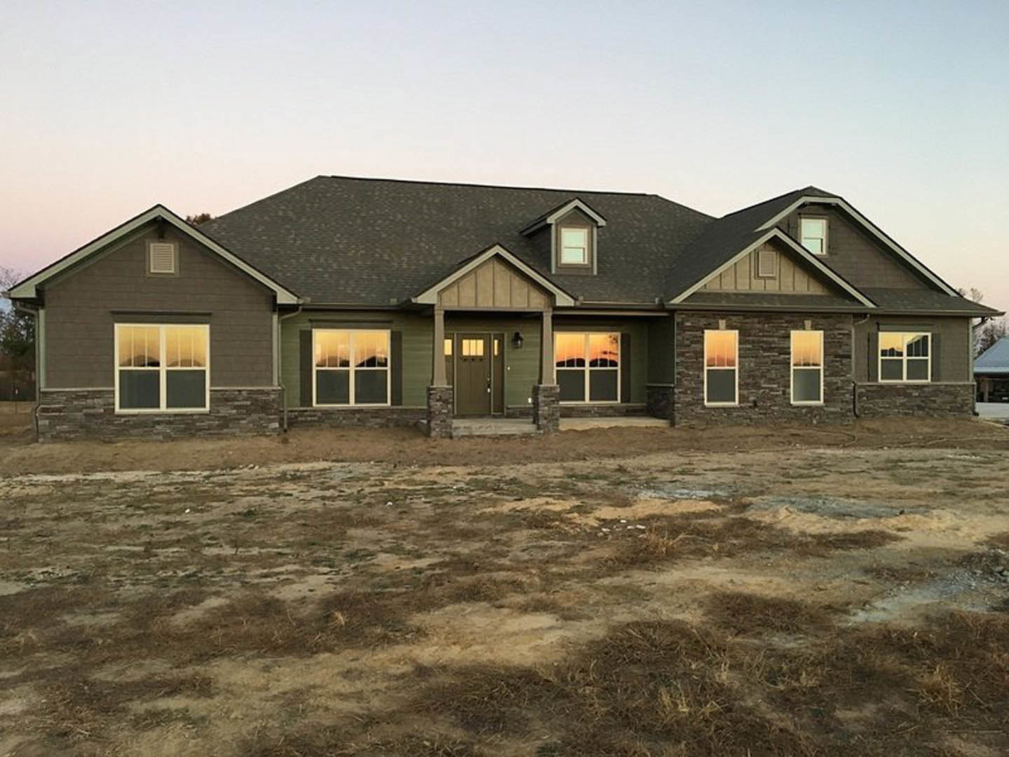 Two-story house with green front door, white-framed windows, gray siding, and expansive grassy yard with scattered rocks; mountain view visible through window.