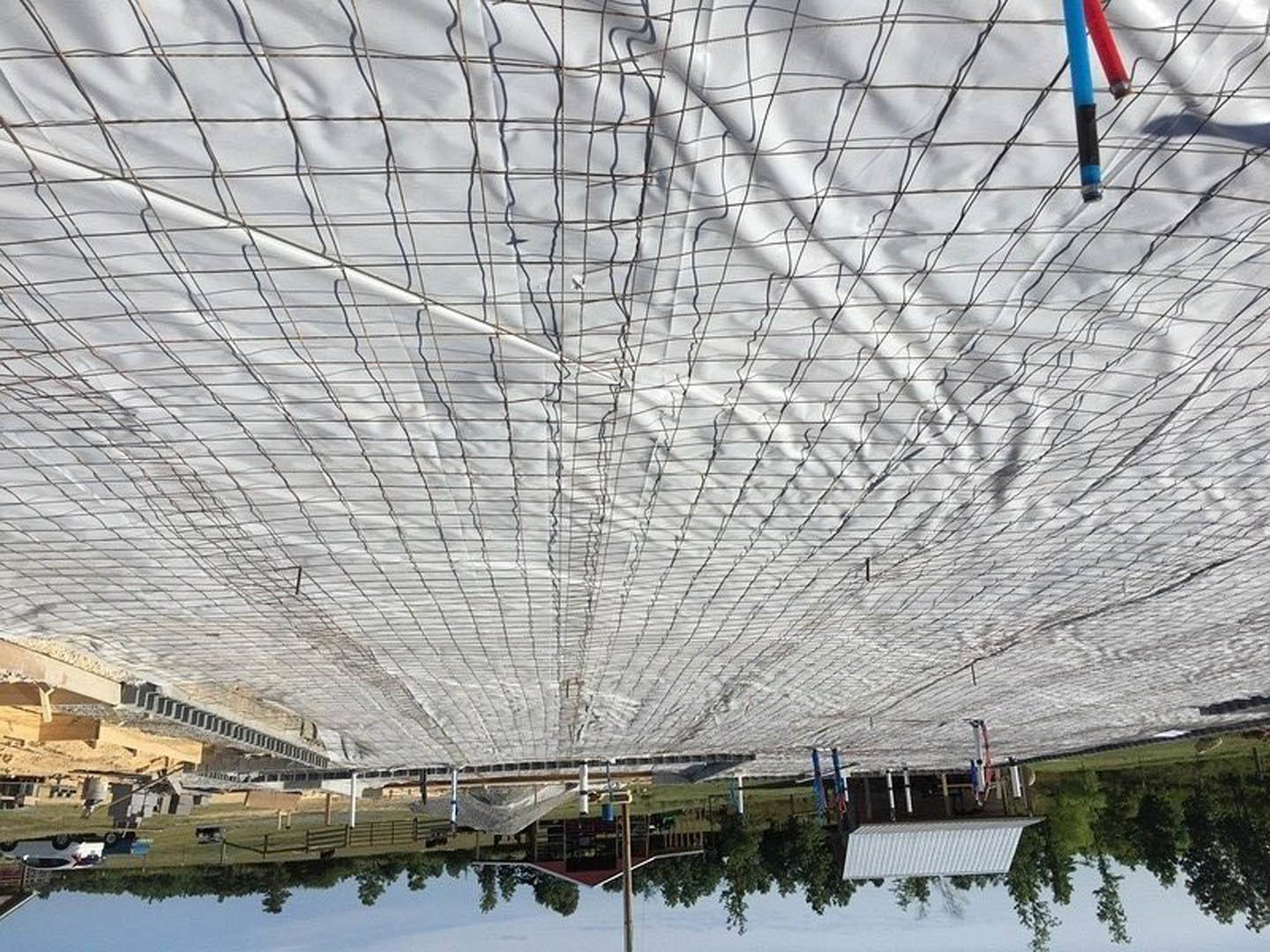 White tarp with blue and red stripes draped over outdoor area near wooden pole, red barn, and blue lake in background